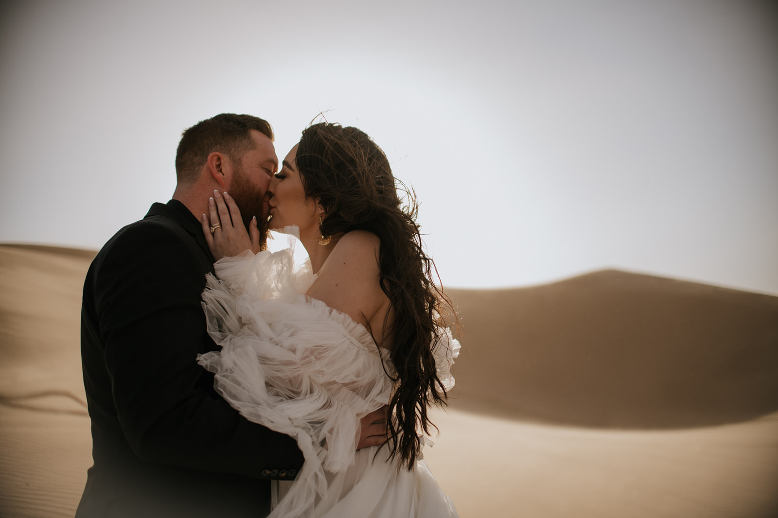 Bride and groom kissing in golden light with sweeping desert dunes behind them during their destination elopement.
