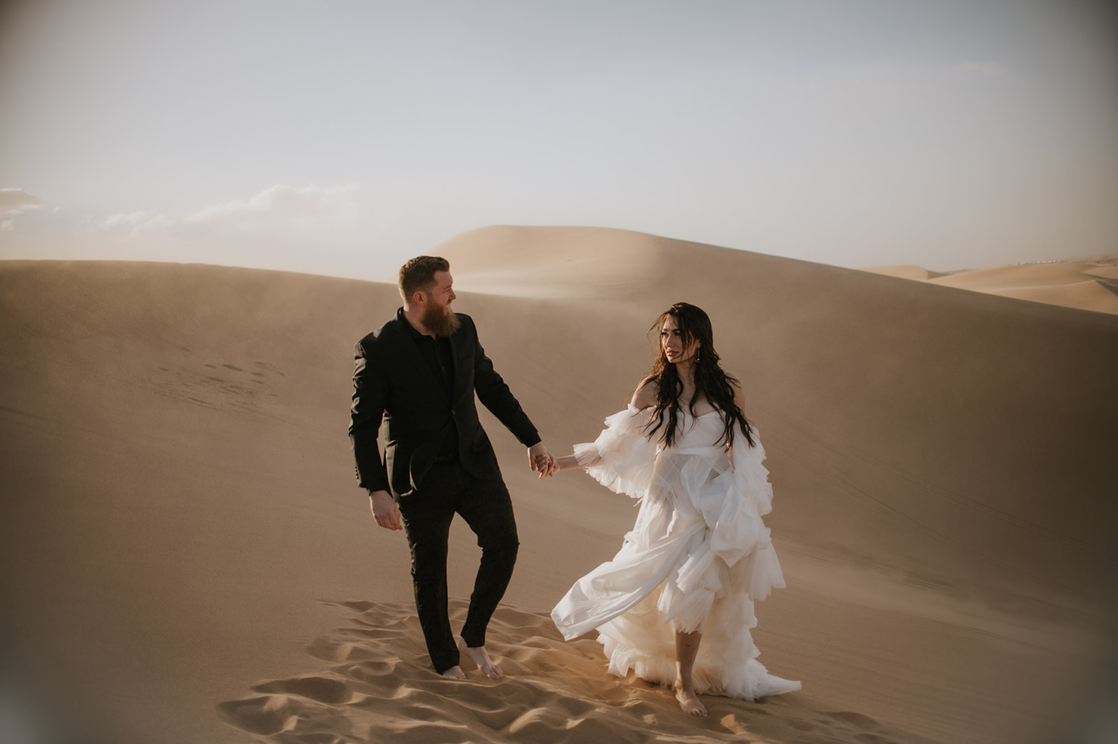 Couple walking barefoot across golden desert dunes while holding hands during their destination elopement.
