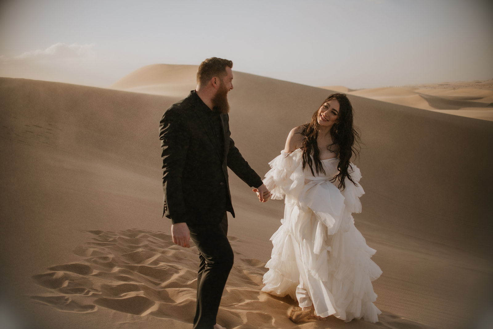 Couple holding hands and walking barefoot through sweeping desert sand dunes at sunset during an intimate destination elopement.
