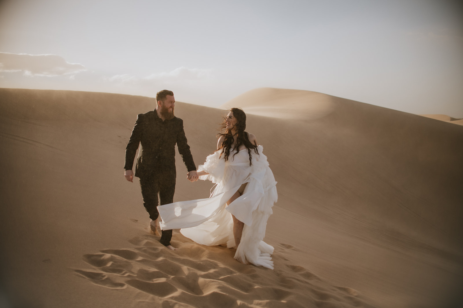 Bride and groom laughing together while walking barefoot through the desert during their destination elopement.
