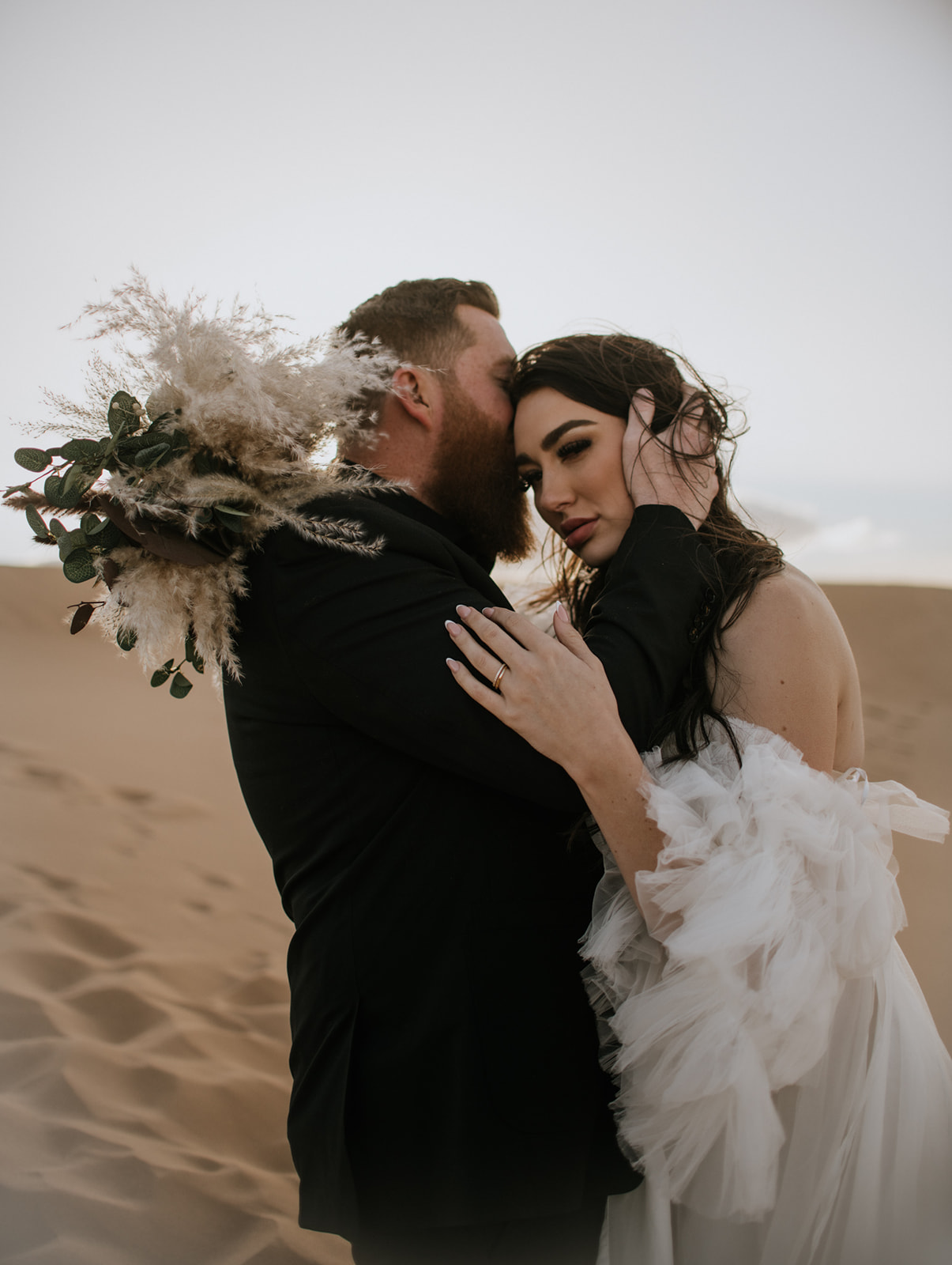 Groom holding the bride close in the desert while she carries a dried pampas bouquet during their destination elopement.
