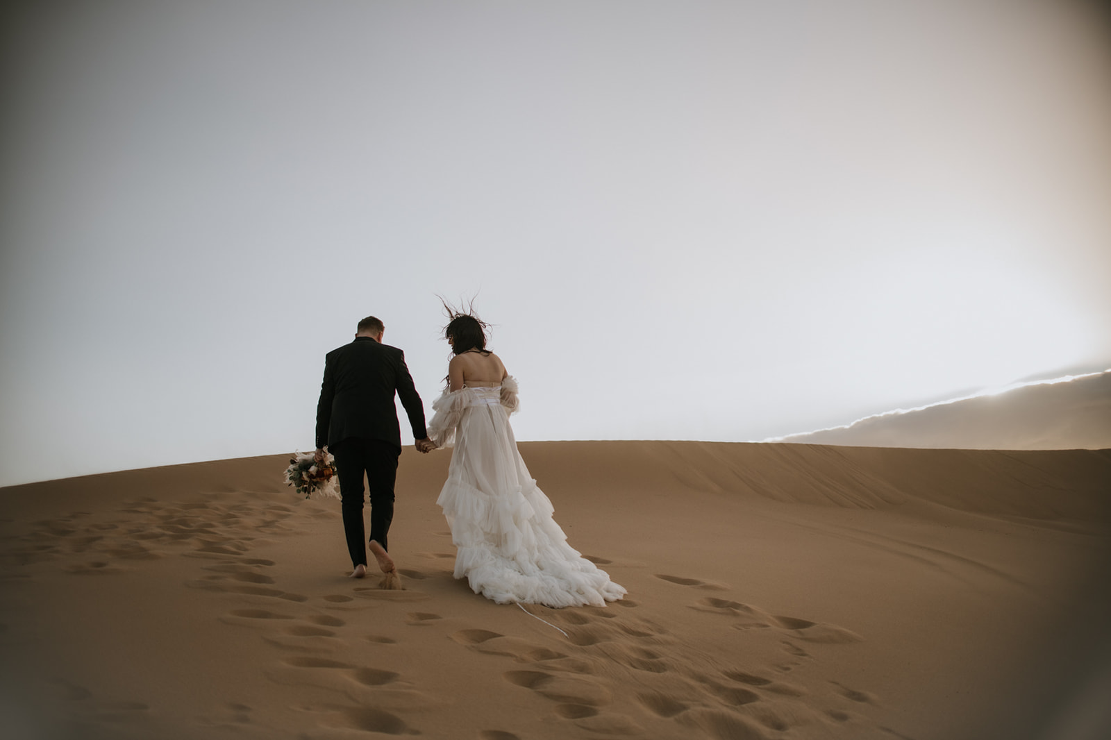 Couple walking hand in hand up a sweeping sand dune during a quiet desert destination elopement.
