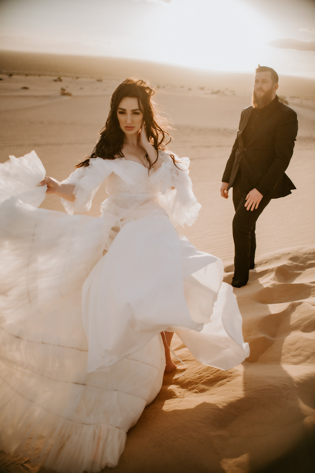 Wind catching the bride’s flowing gown as she walks across sunlit sand dunes with the groom following behind.
