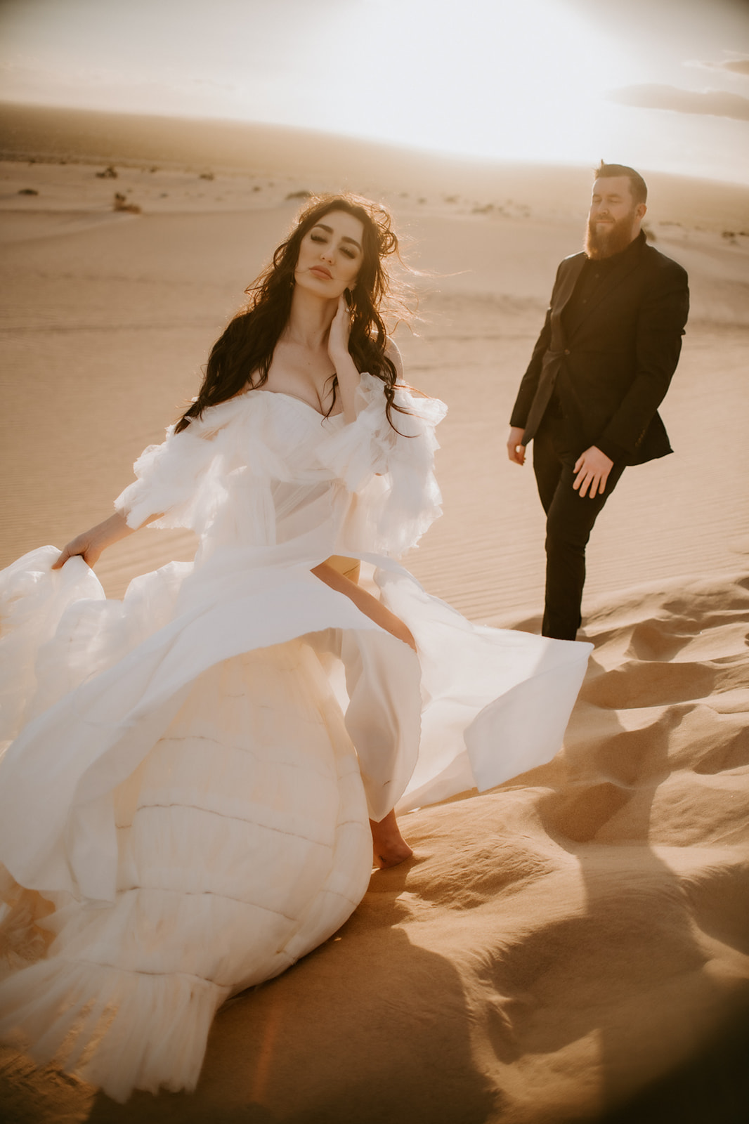 Wind catching the bride’s layered dress as she walks through sunlit sand dunes with the groom following behind.
