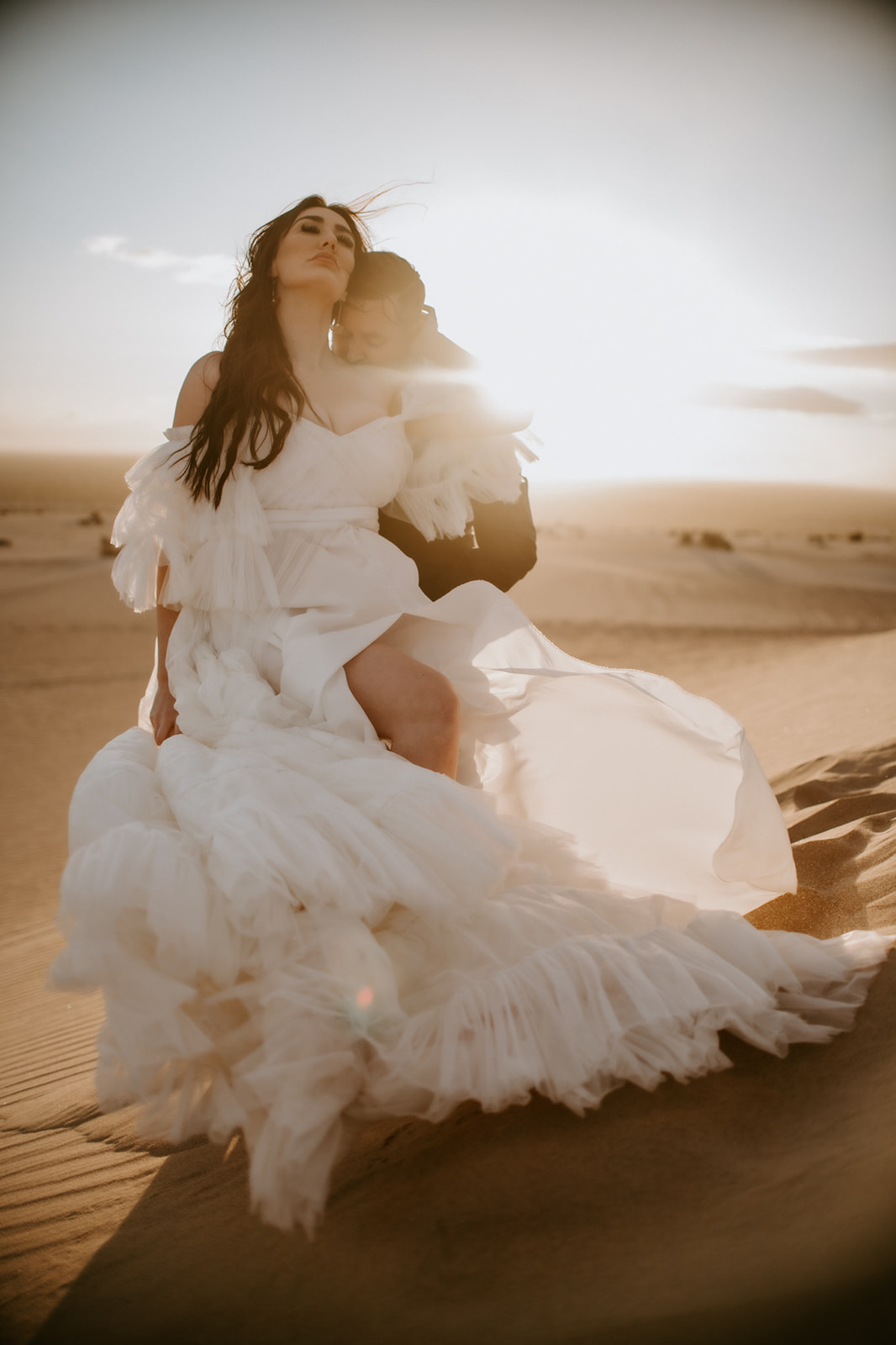 Groom embracing the bride from behind as sunlight glows through her flowing dress on a desert dune.
