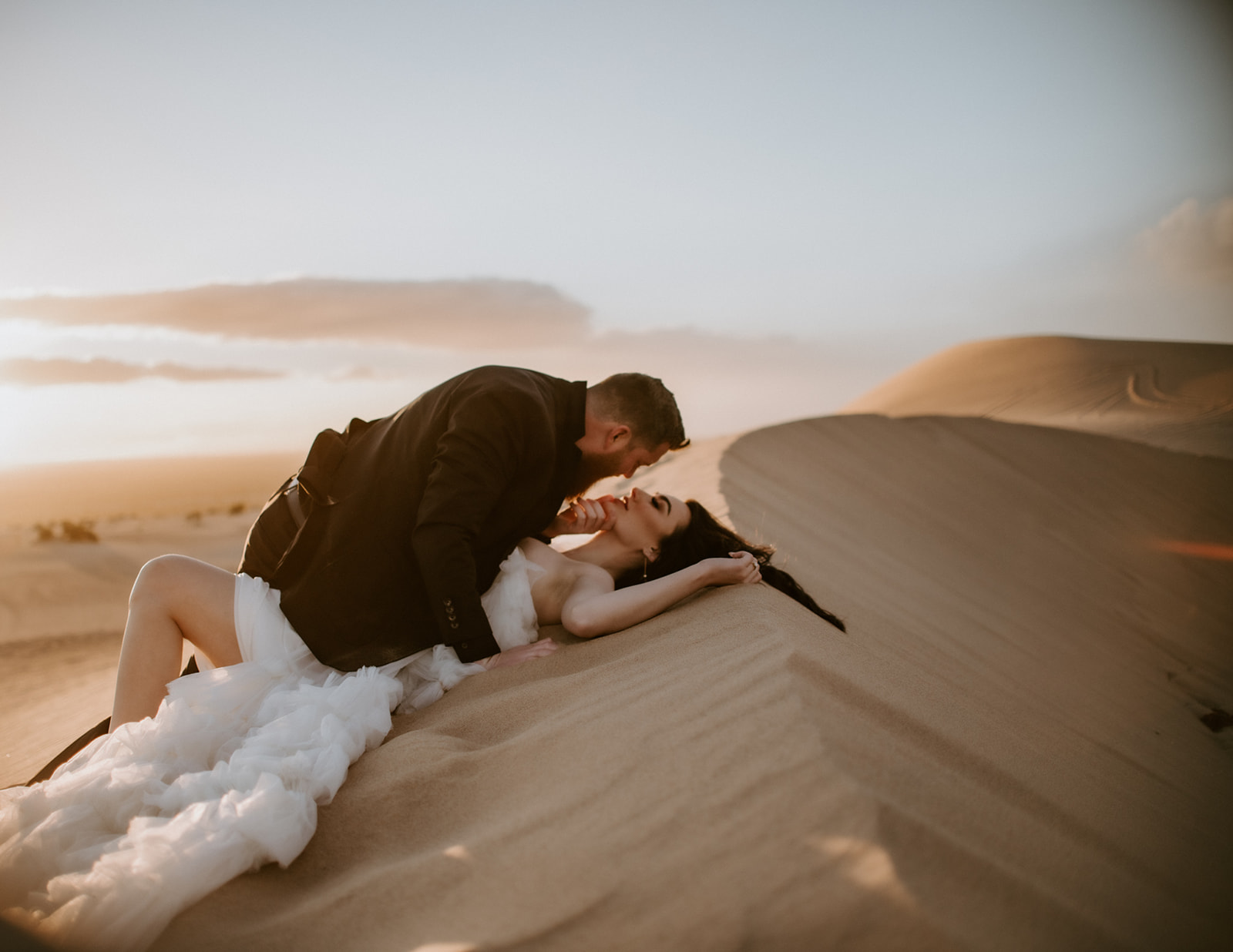 Groom leaning over the bride as she reclines on a warm sand dune during an intimate desert destination elopement.
