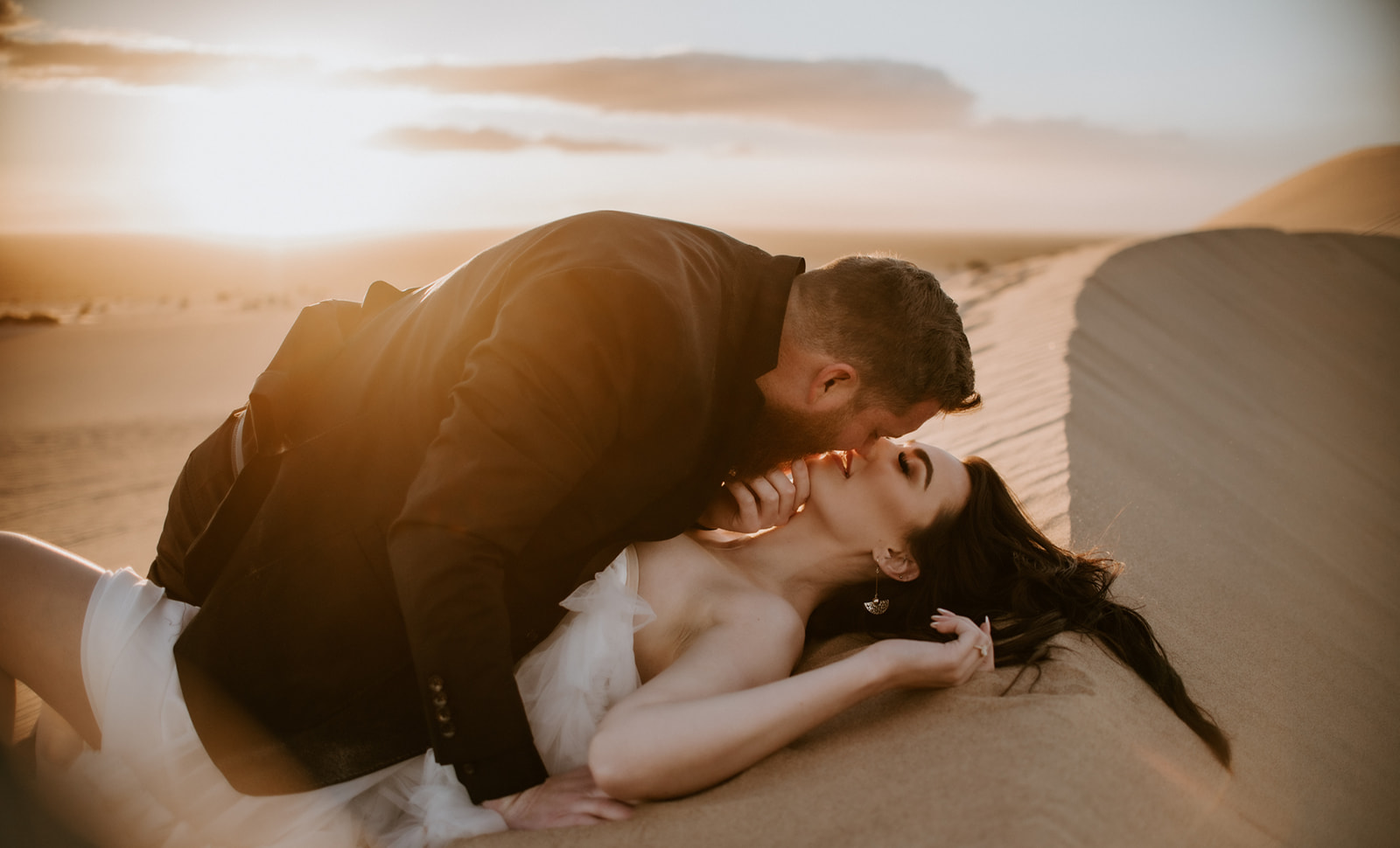 Groom kissing the bride as she reclines in warm desert sand during their destination elopement.
