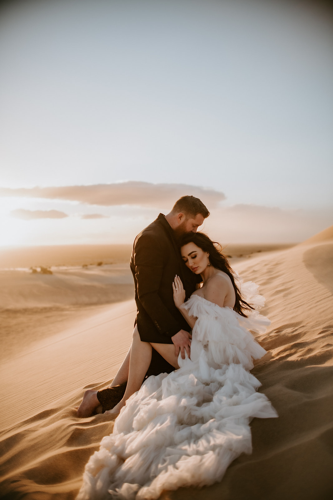 Bride resting against the groom as they sit together on a glowing desert dune during their destination elopement at golden hour.
