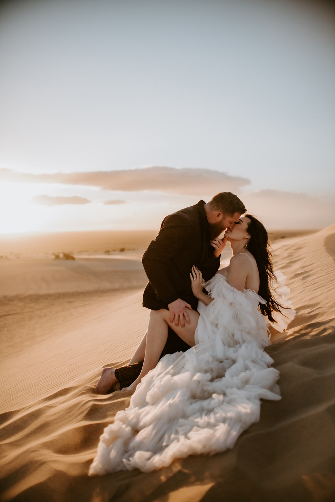 Bride and groom sharing a kiss while sitting together on a warm desert dune during their destination elopement at golden hour.
