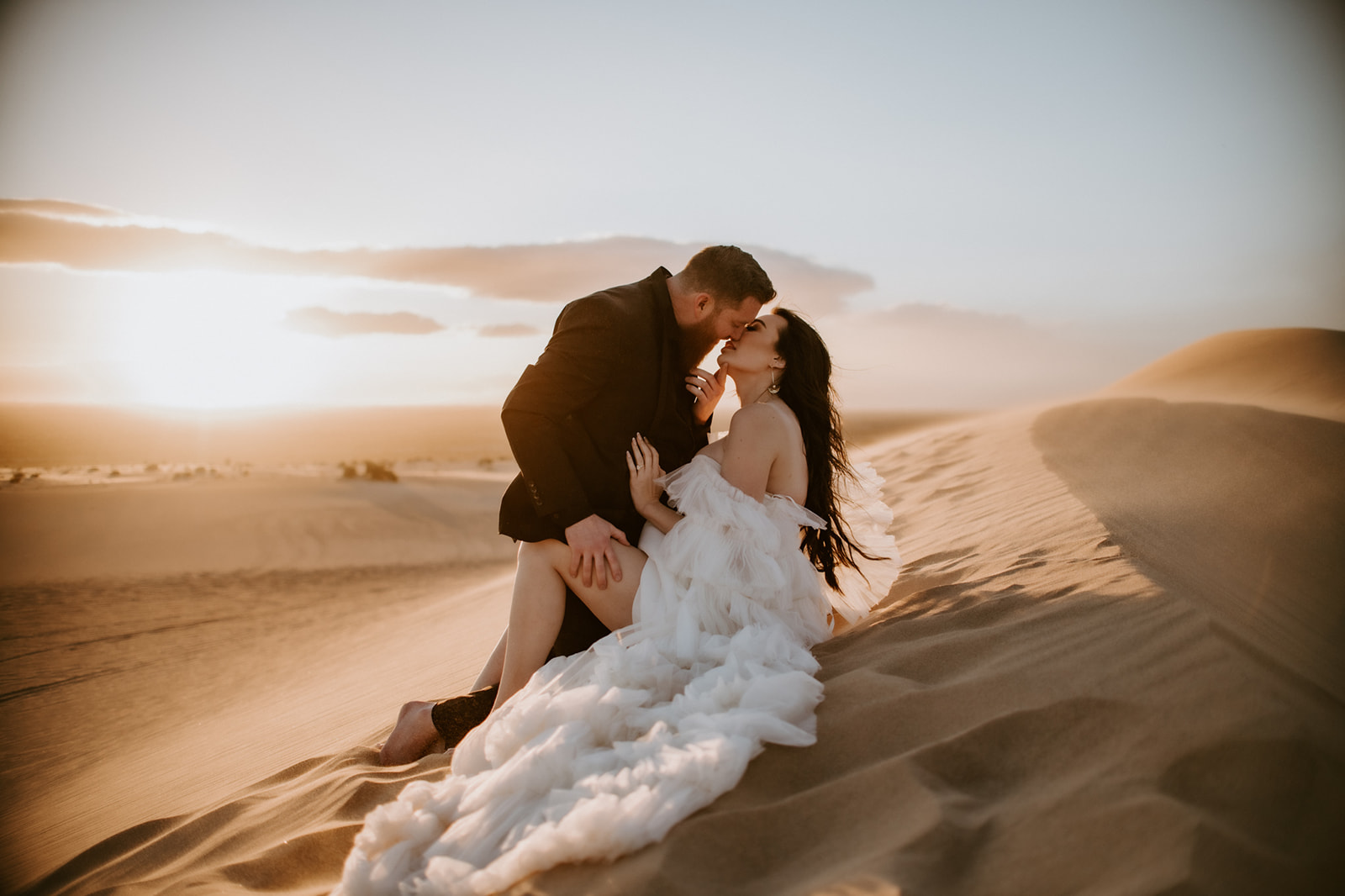 Groom leaning in to kiss the bride while they sit together on a glowing sand dune during their destination elopement.

