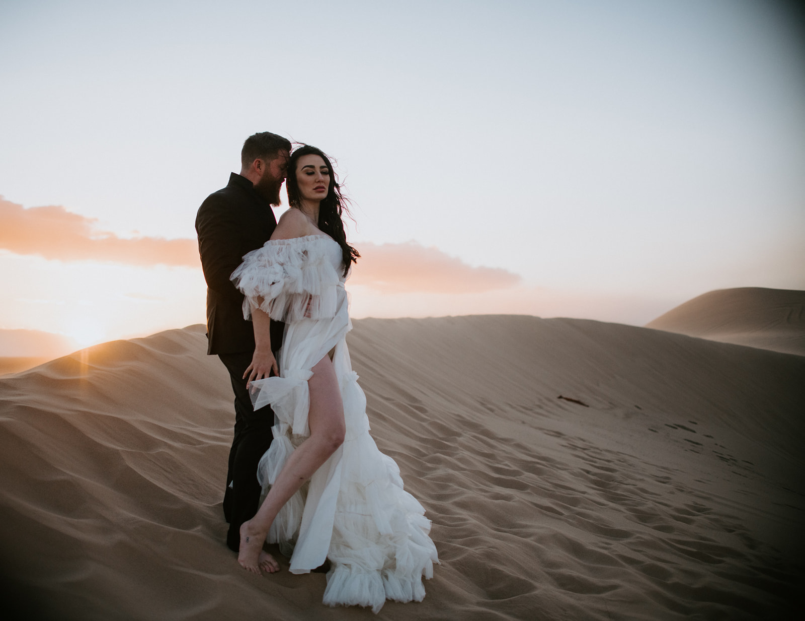 Groom standing behind the bride on a wind-swept sand dune at sunset during an intimate desert destination elopement.
