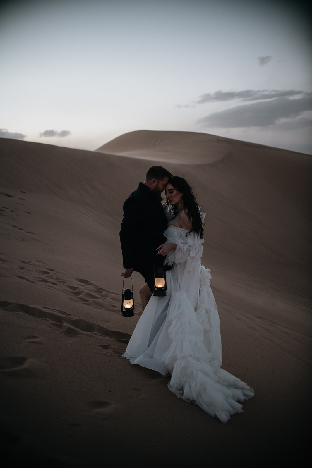 Couple walking slowly through desert sand with lantern light glowing as evening falls during their destination elopement.
