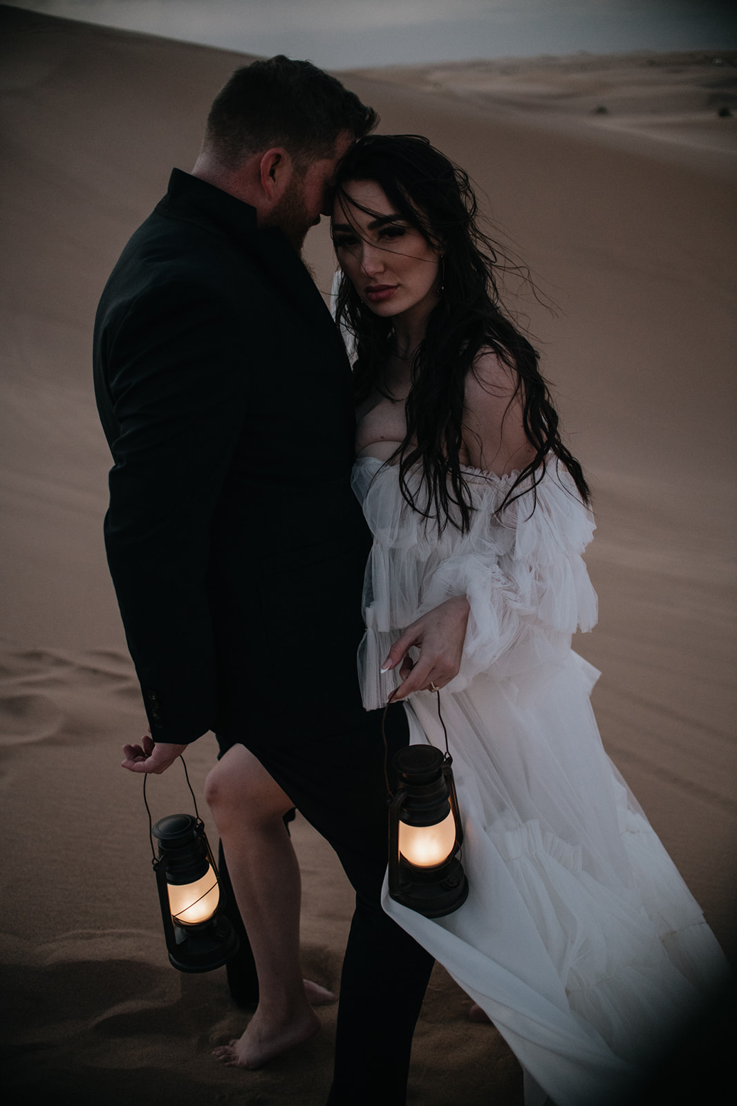 Bride and groom walking through quiet desert dunes at dusk while holding lanterns during a moody destination elopement moment.
