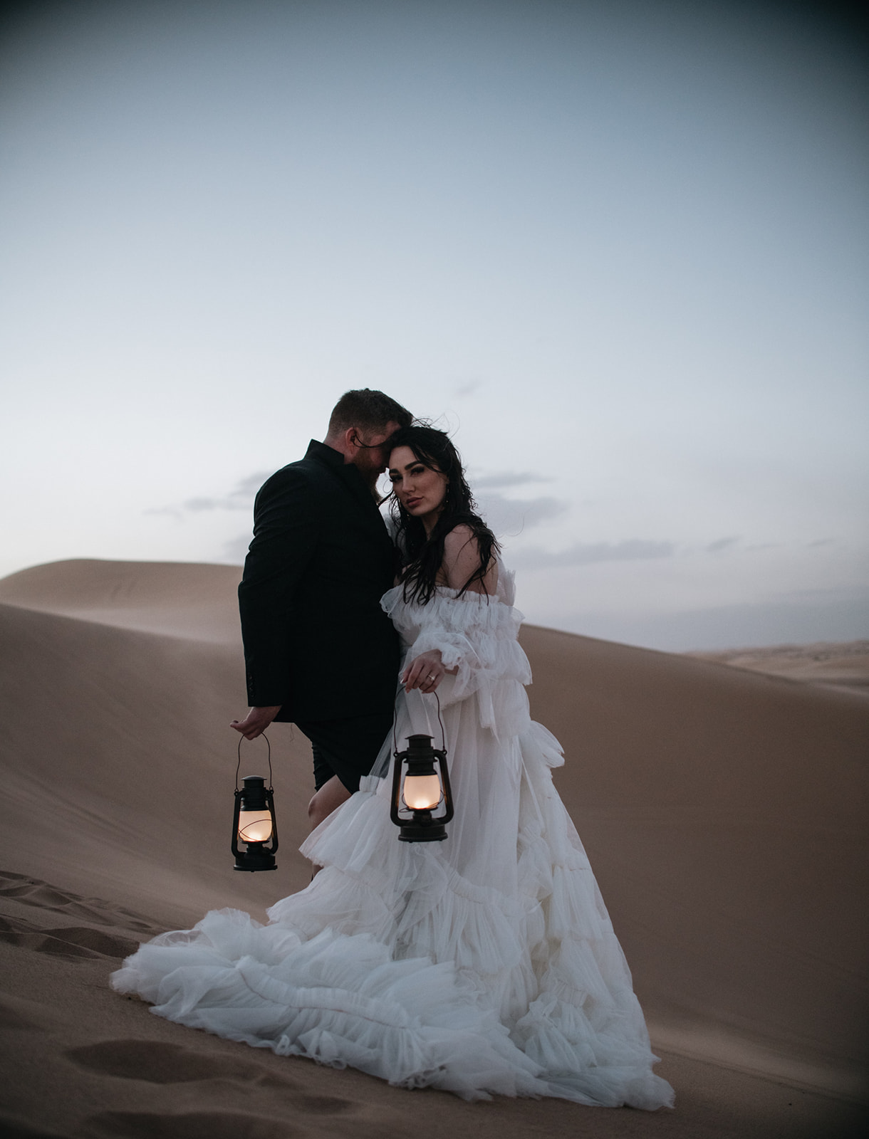 Bride and groom standing close together on a wind-swept dune holding lanterns at twilight.
