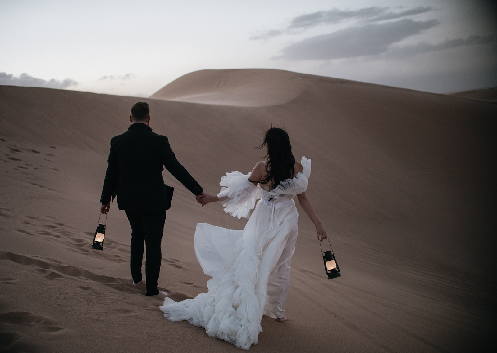 Bride and groom walking across rippled sand dunes holding lanterns during a moody desert destination elopement.
