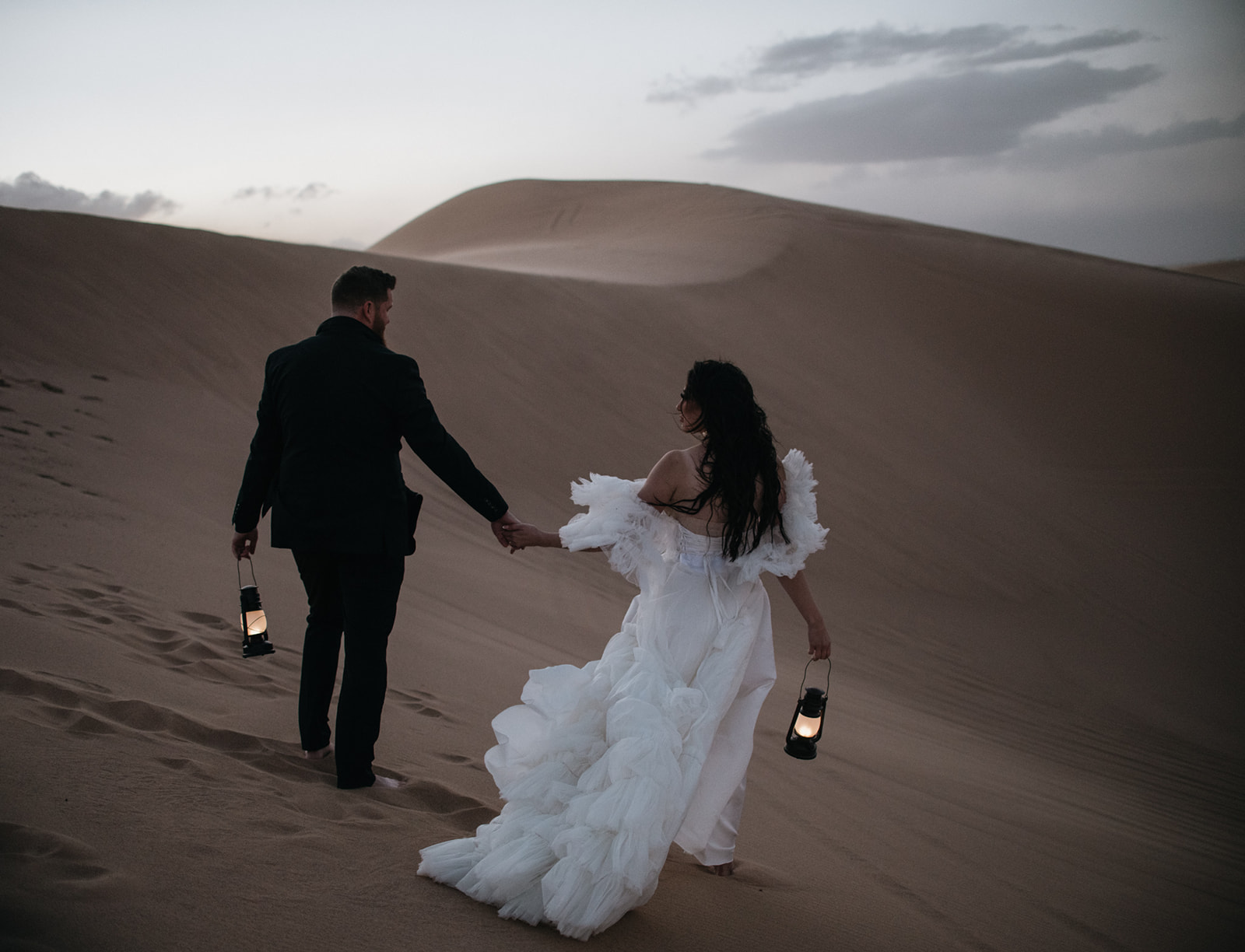 Bride and groom walking across desert dunes at dusk holding lanterns during their destination elopement.
