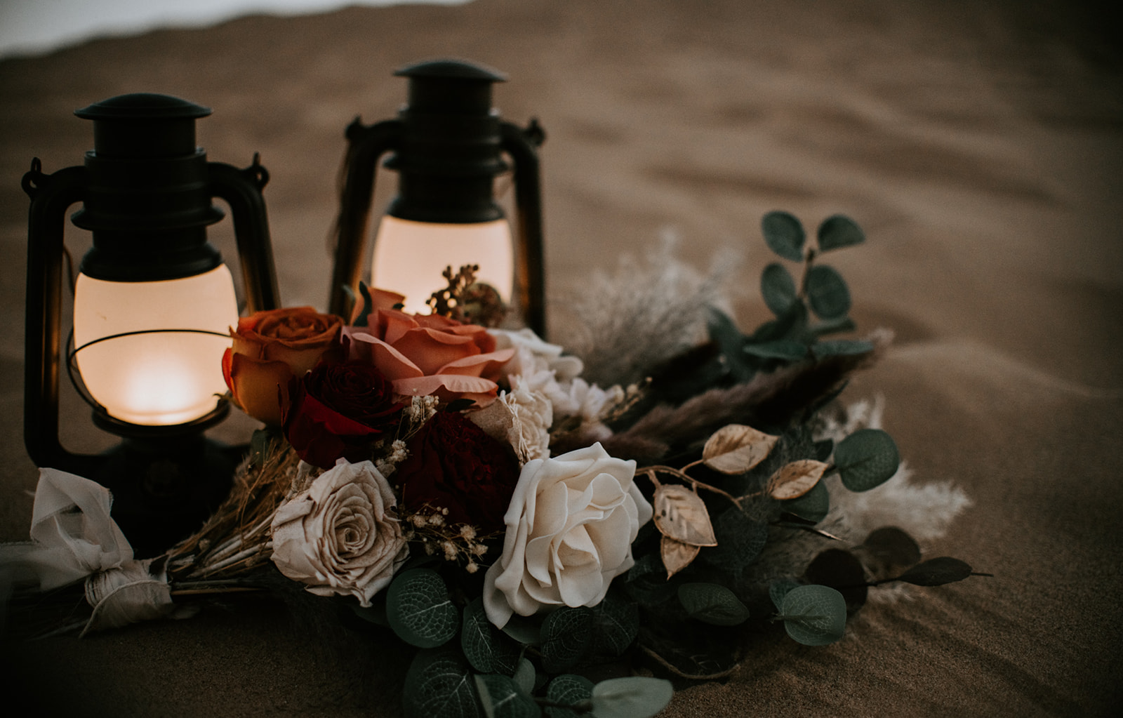 Desert elopement bouquet with roses, pampas grass, and greenery beside glowing lanterns in the sand.
