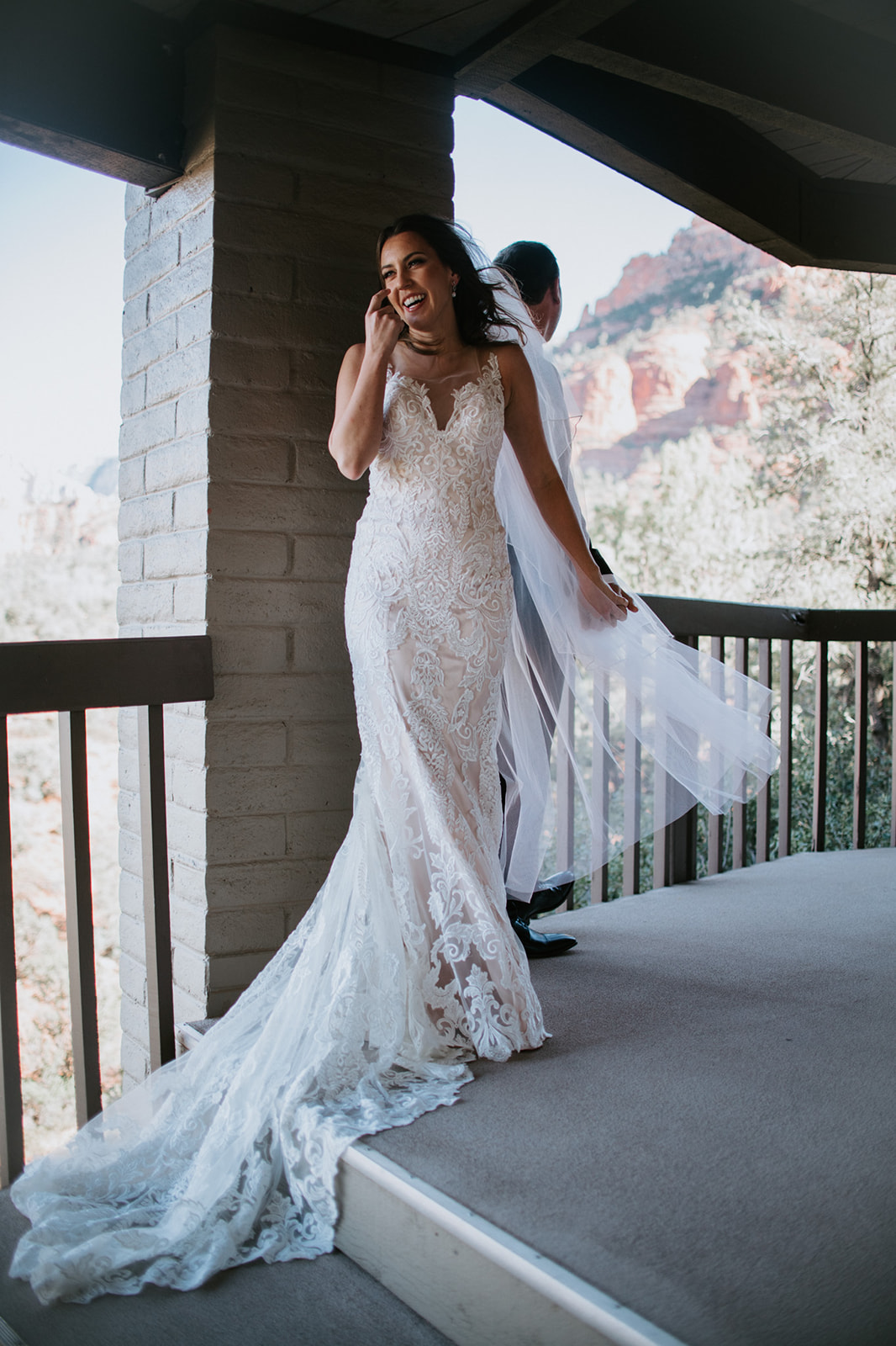 Bride laughing on a balcony with veil moving in the breeze and red rock landscape behind her
