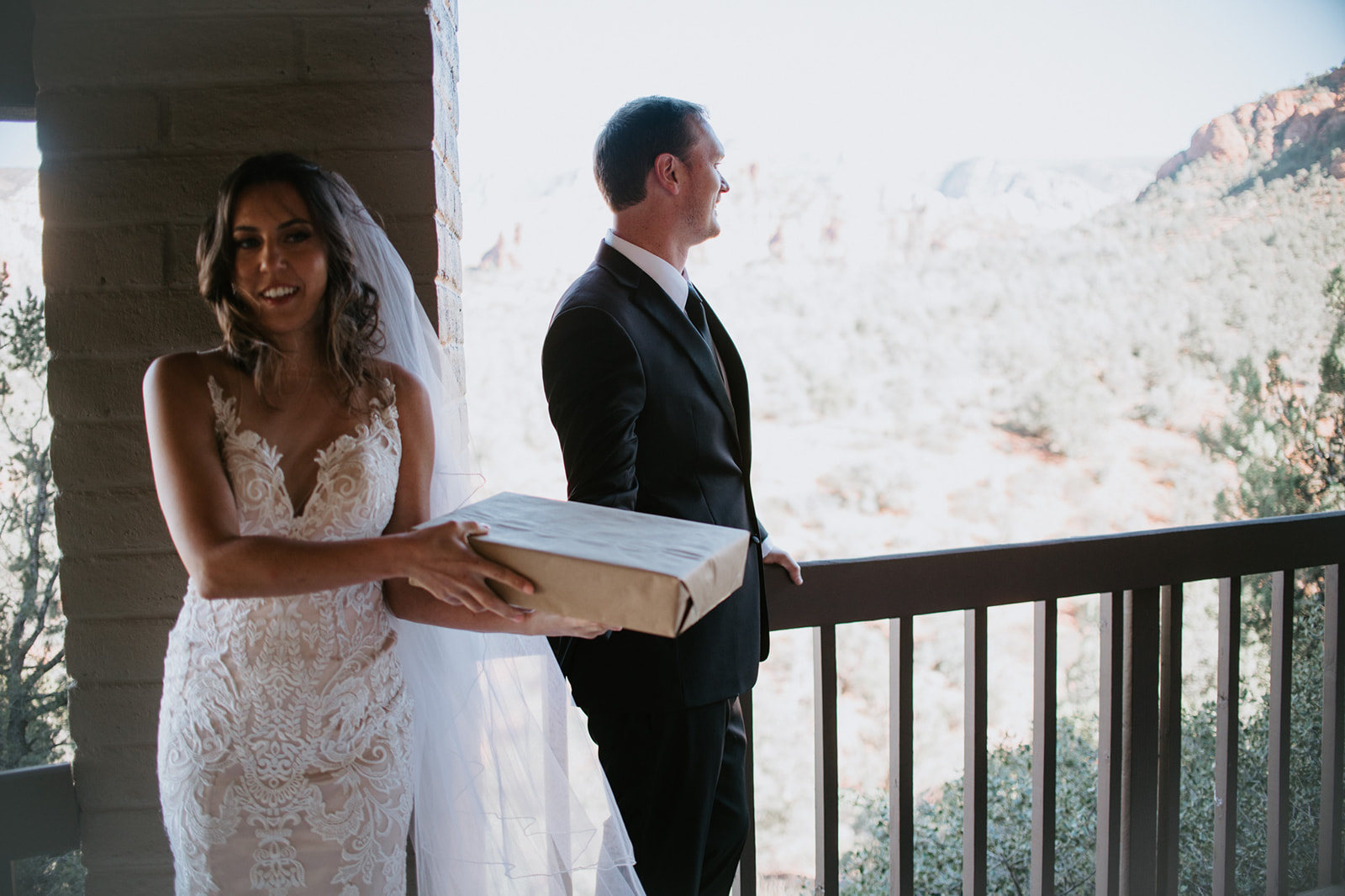 Bride holding a wrapped gift on a balcony while groom looks out over the desert landscape
