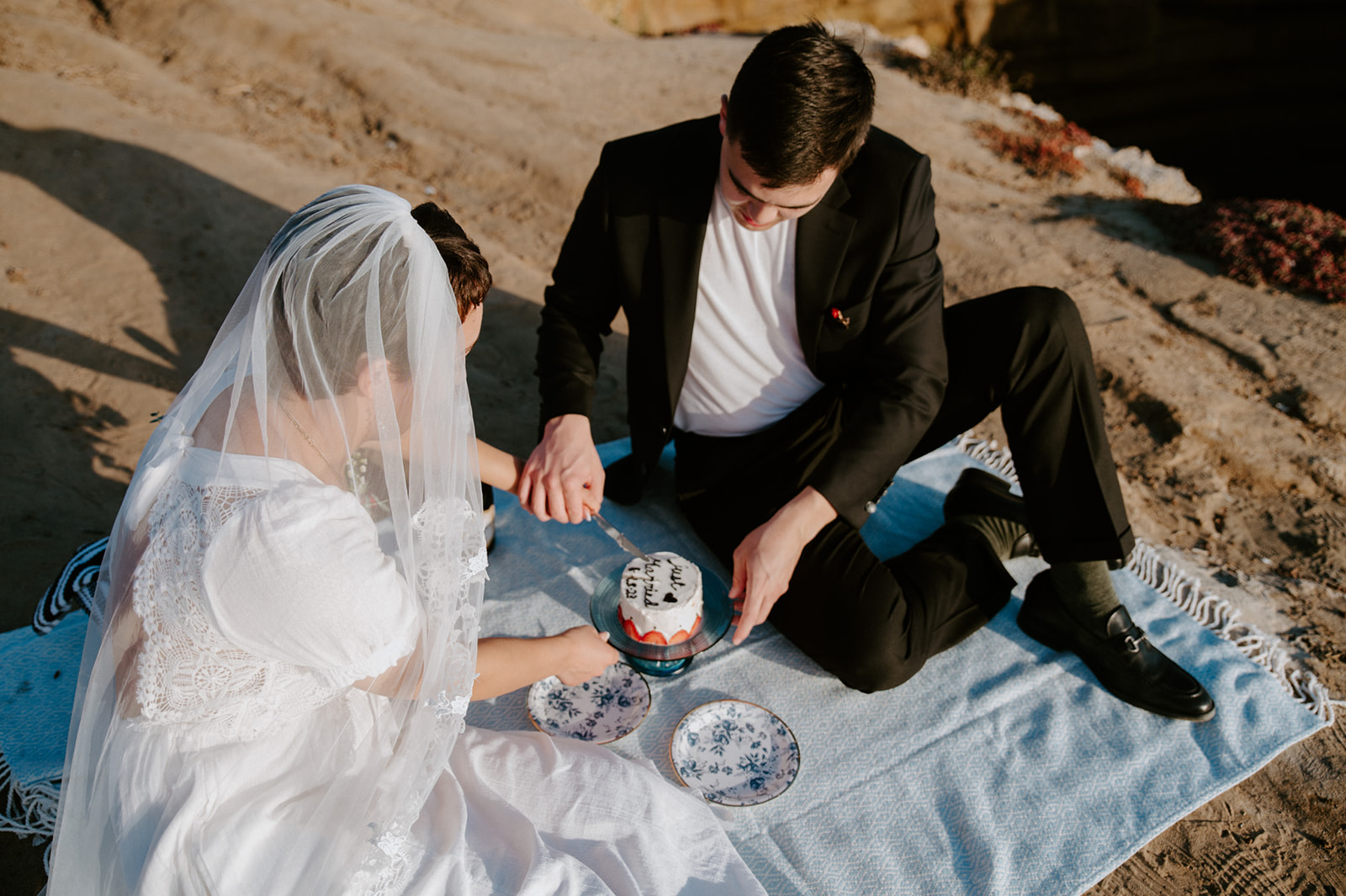 Couple sitting on a blanket cutting their small wedding cake together, a relaxed picnic moment from their elopement checklist.

