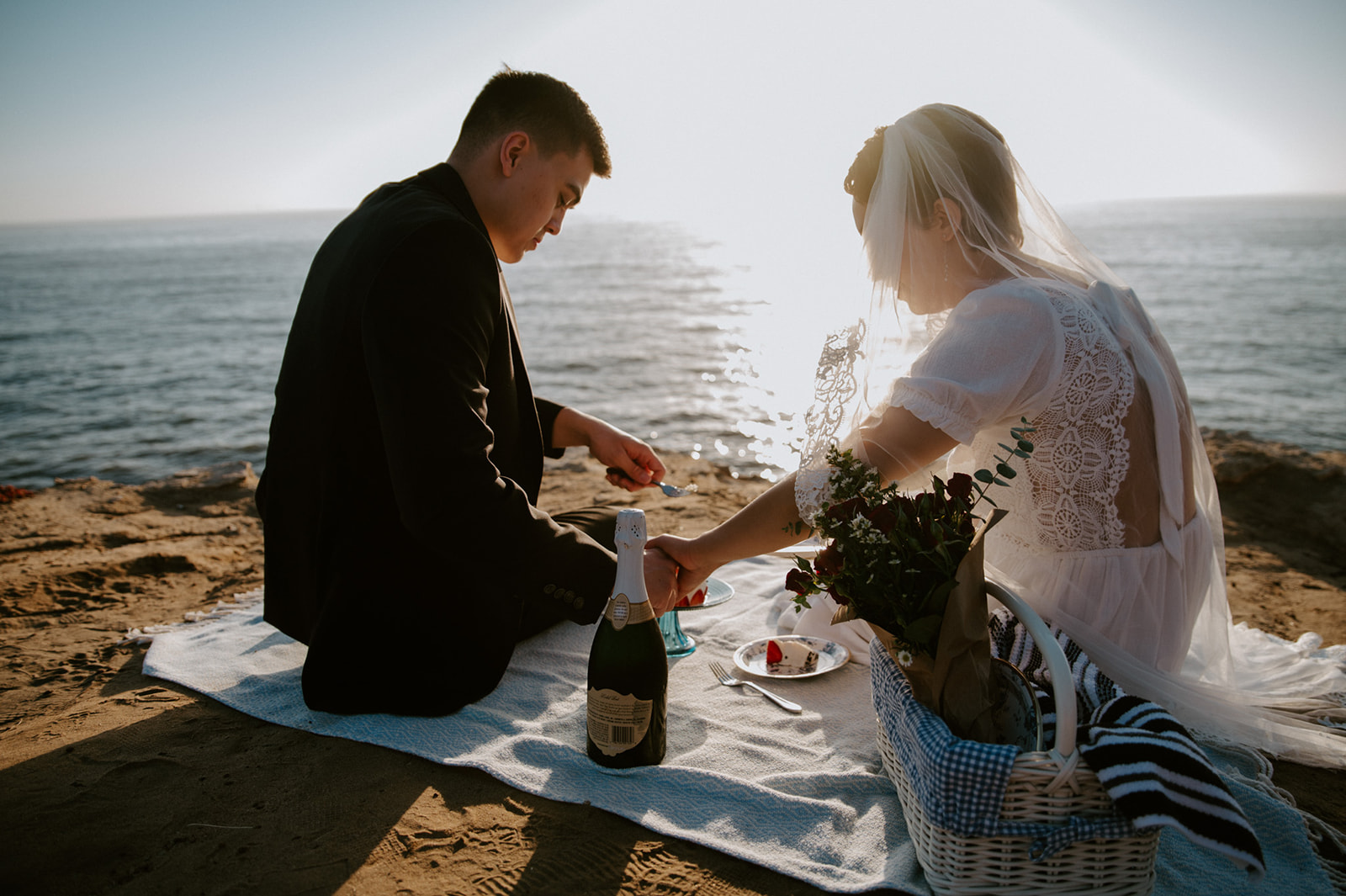 Couple sitting together on a blanket by the ocean cutting into their cake, a relaxed and intentional moment from their elopement checklist.
