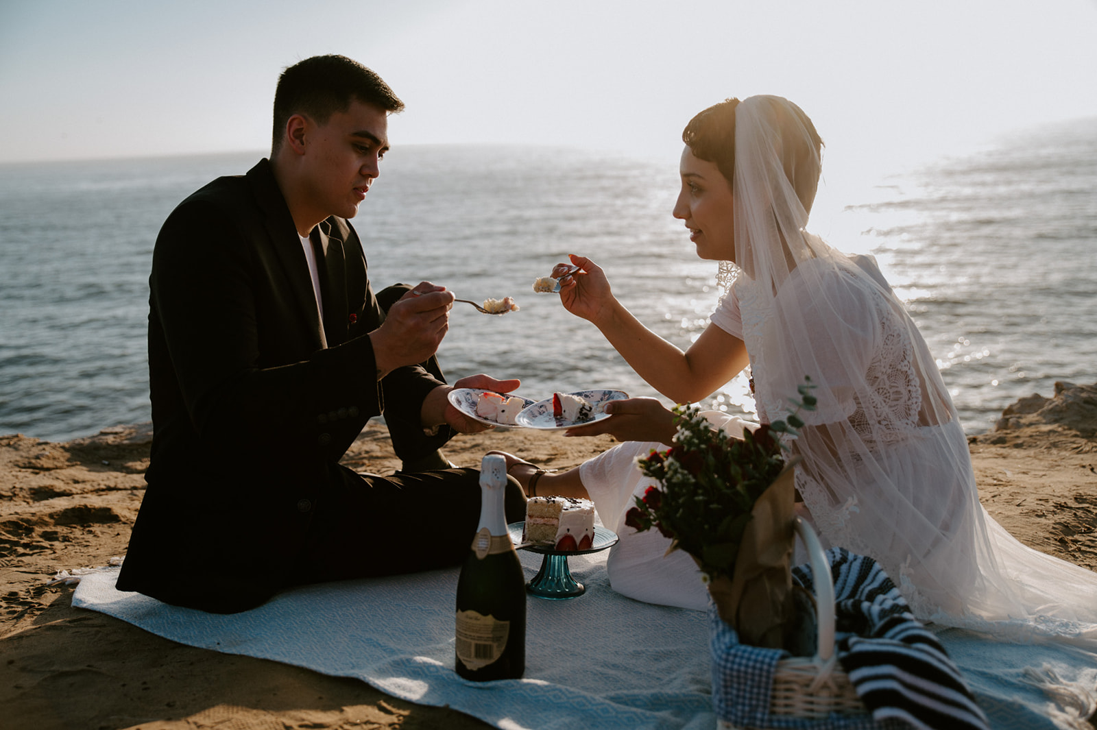 Couple feeding each other cake on a picnic blanket by the ocean, a playful and personal detail from their elopement checklist.
