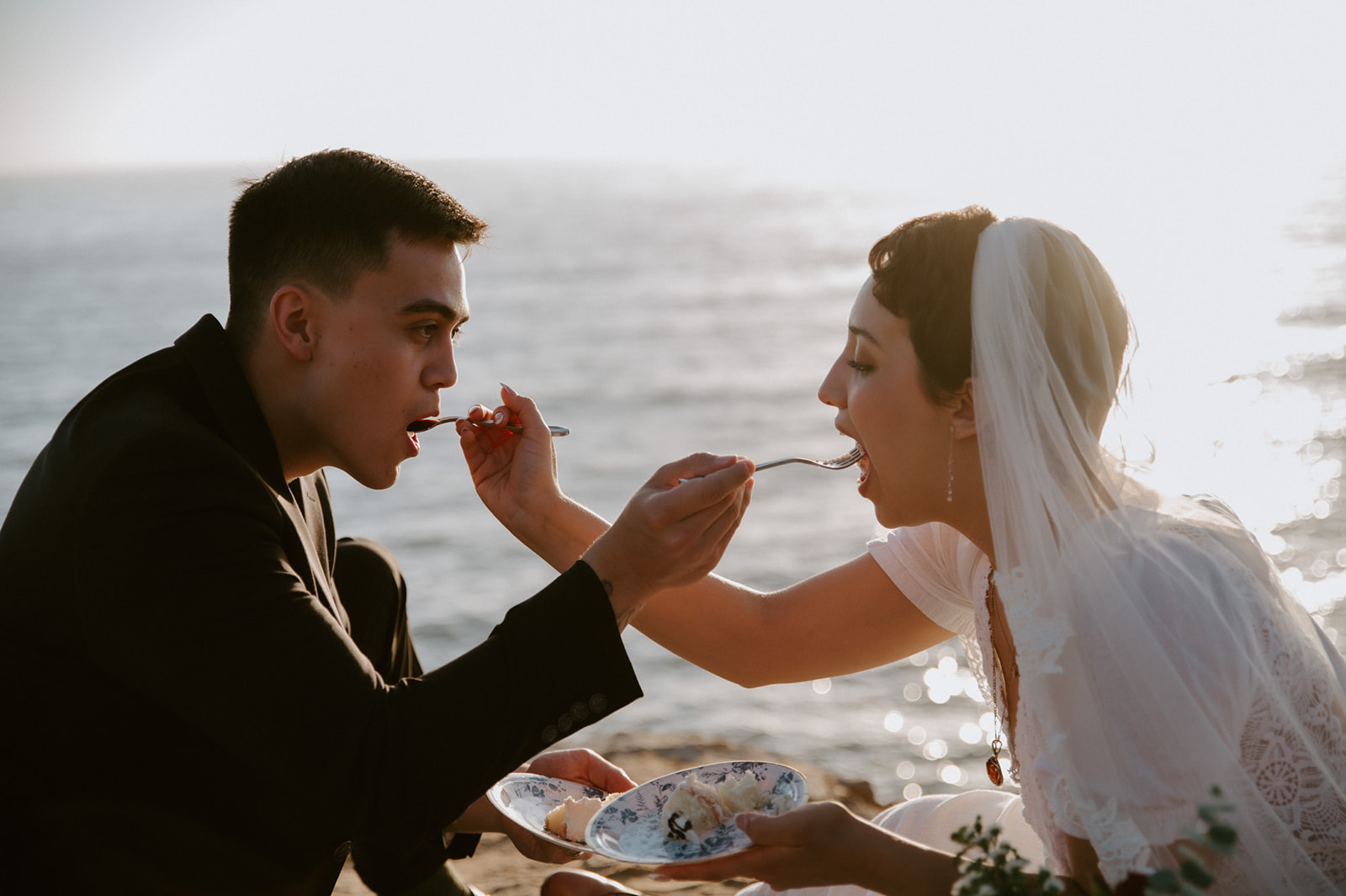 Couple feeding each other cake by the ocean at sunset, a playful and intimate elopement experience.

