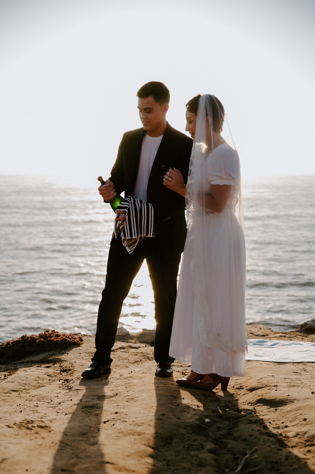 Couple opening a champagne bottle together on a coastal cliff at sunset, celebrating a just-married moment from their elopement checklist.

