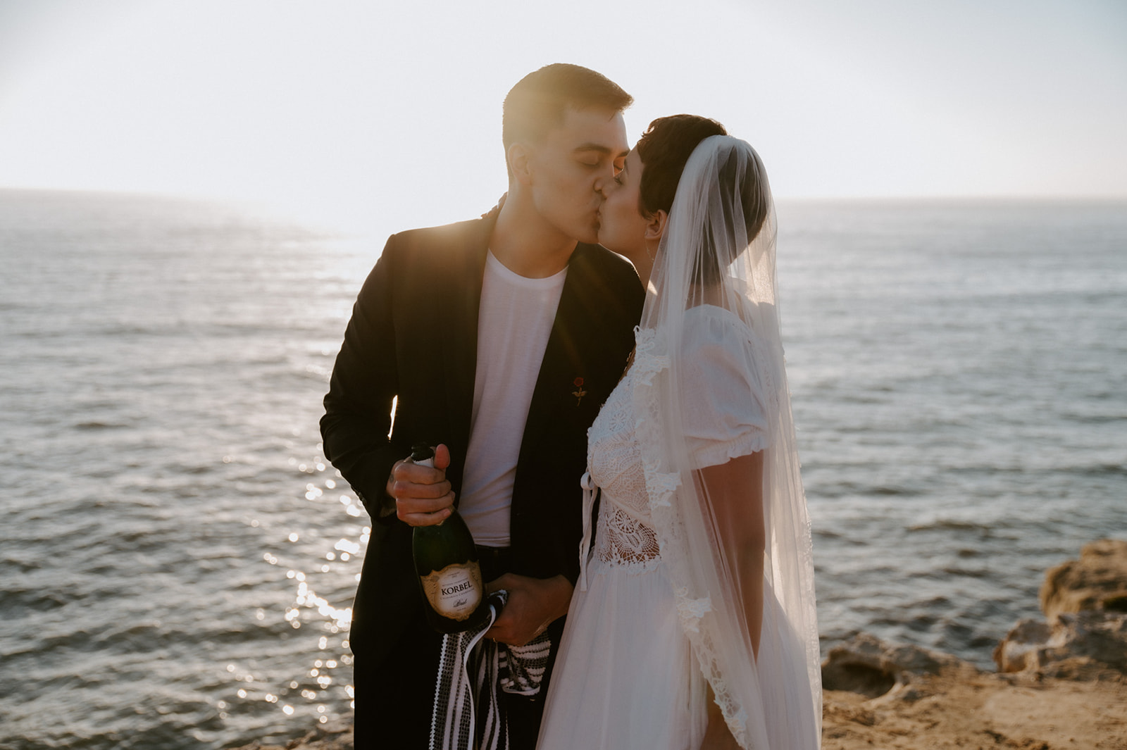 Couple sharing a kiss while holding a champagne bottle at sunset, a celebratory moment during their elopement.
