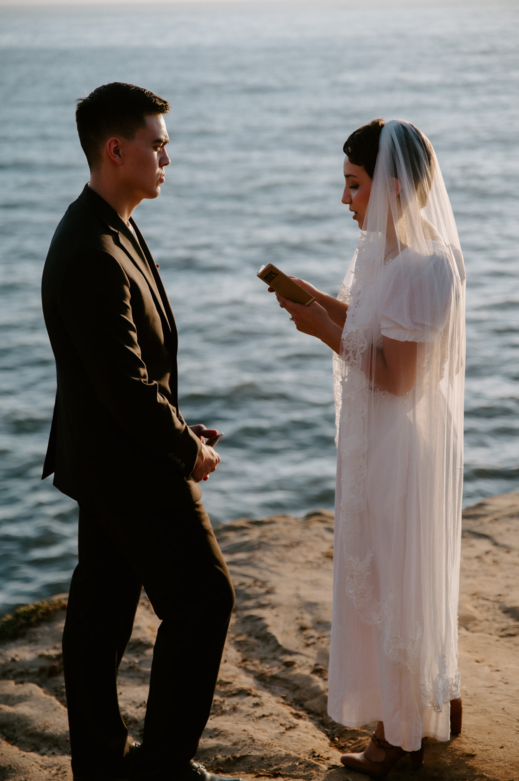 Bride reading her vows during a seaside ceremony, referencing a handwritten notebook from their intentional elopement checklist planning.
