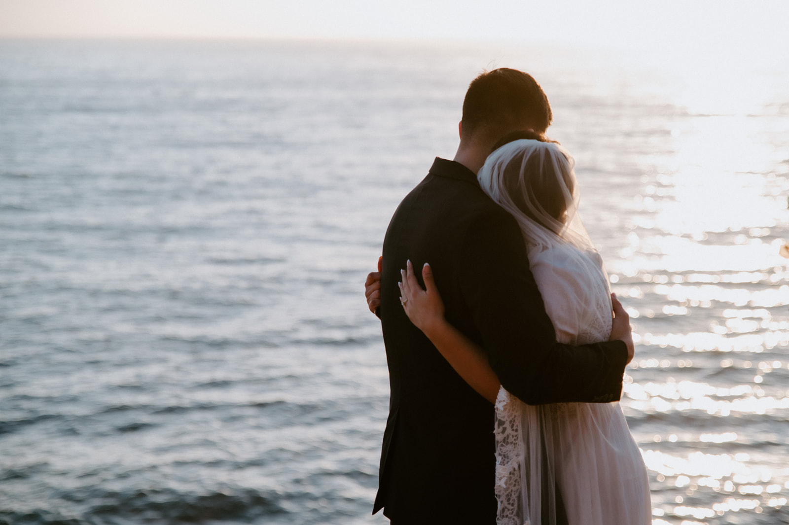 Couple embracing from behind while watching the sun reflect on the ocean, a quiet just-married moment from their elopement checklist.
