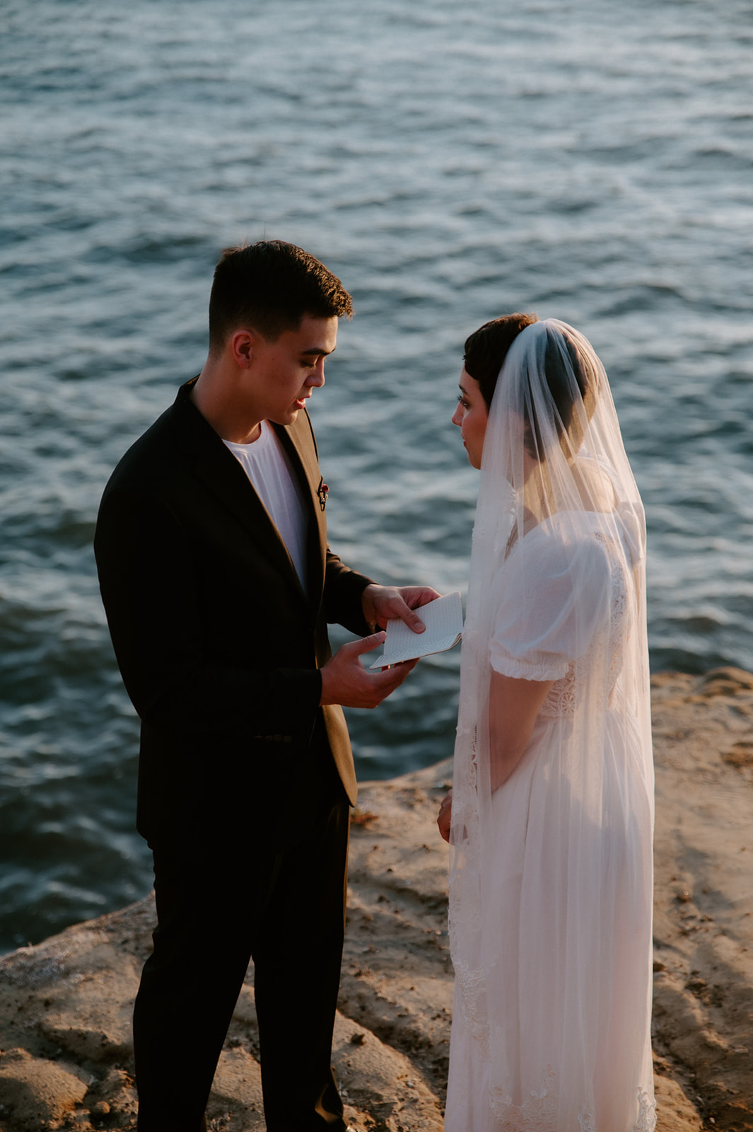Groom reading vows from a notebook to his partner on a cliffside overlooking the ocean, part of a meaningful elopement checklist experience.
