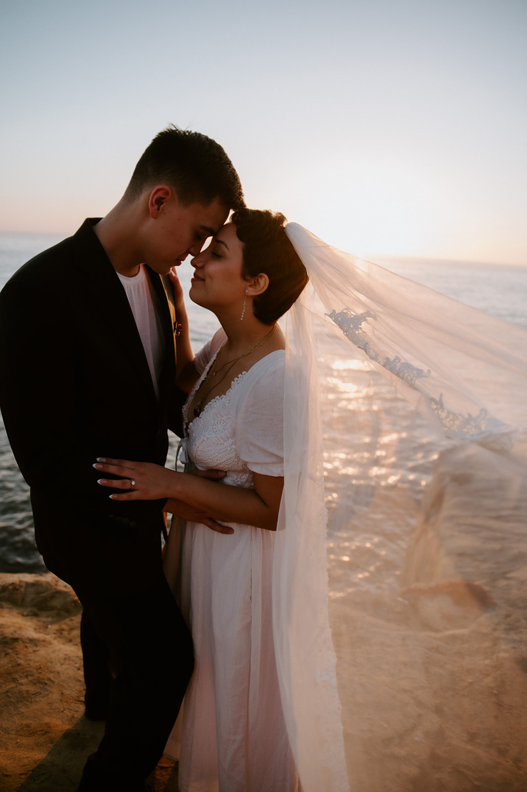 Bride and groom standing together on a coastal cliff with the bride’s veil blowing in the wind, a candid moment from their elopement checklist day.
