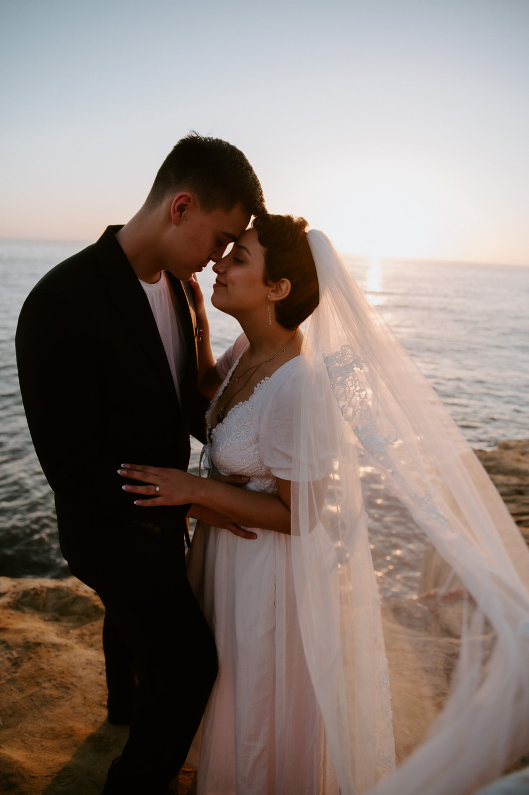 Couple standing forehead to forehead on a coastal cliff at sunset, the bride’s veil catching the ocean breeze during their elopement checklist day.
