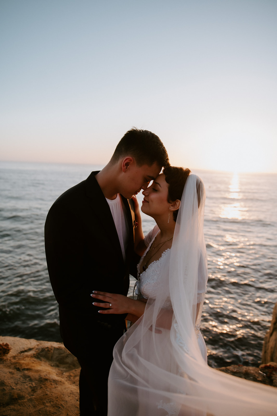 Couple leaning into each other with foreheads touching as the sun sets over the ocean, a quiet and emotional elopement moment.
