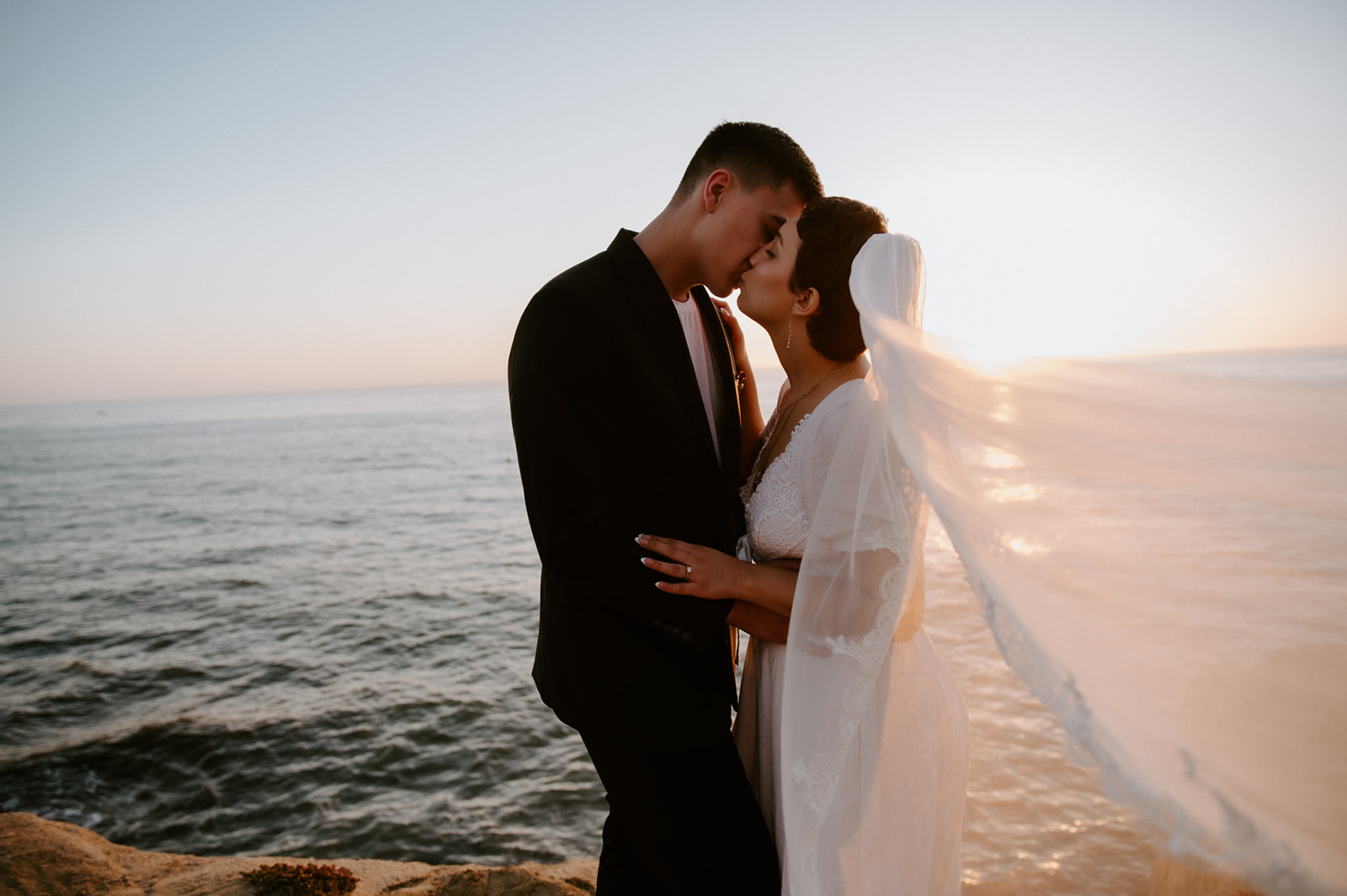 Couple standing close with the bride’s veil flowing in the ocean breeze at sunset, a cinematic moment from their elopement checklist.
