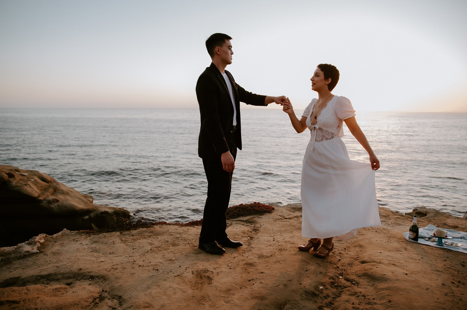 Couple holding hands and dancing along a seaside bluff at golden hour, a quiet picnic setup nearby for their elopement celebration.
