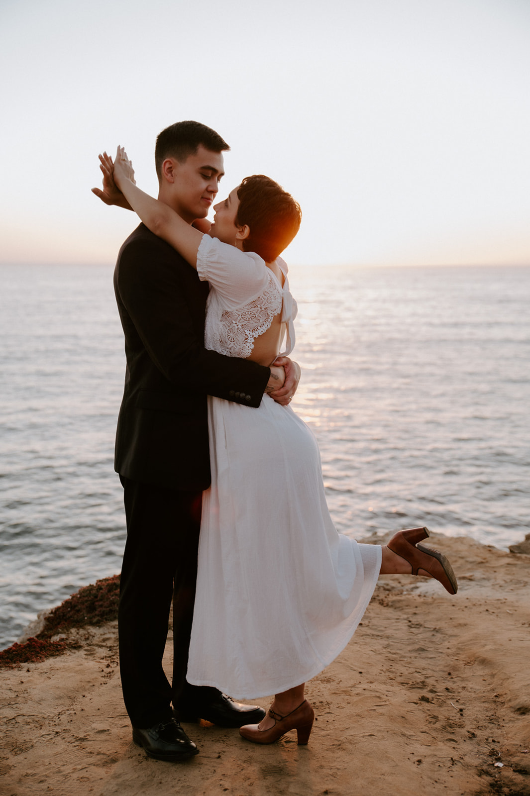 Couple embracing at sunset on a coastal overlook, the bride lifting her leg in a playful and intimate elopement moment.
