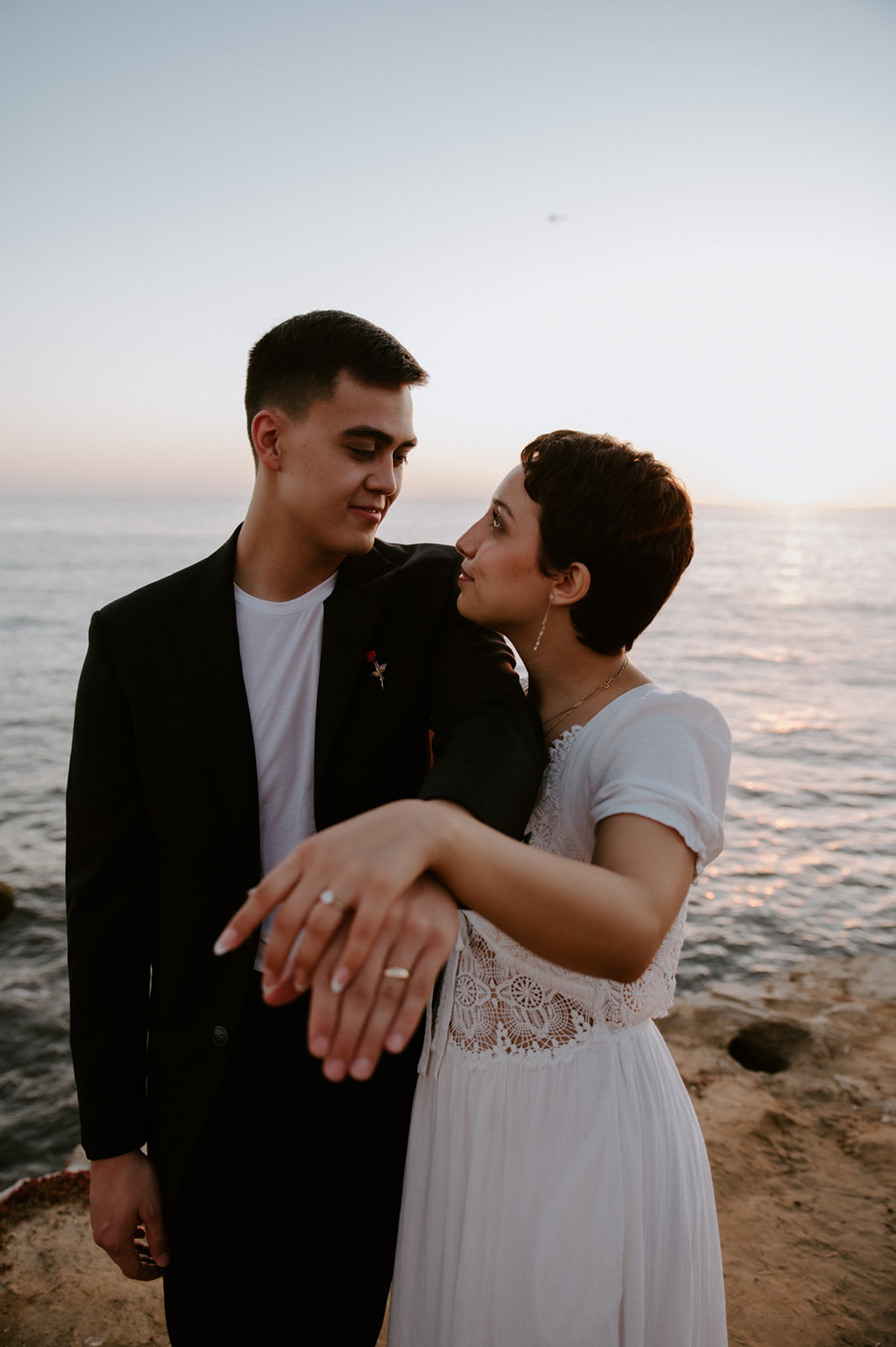 Couple laughing and holding hands near the ocean, showing off wedding rings during a relaxed coastal elopement.
