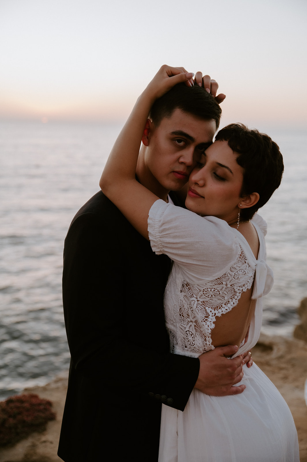 Couple embracing on a coastal cliff at sunset, ocean stretching behind them as the bride rests her head against her partner.
