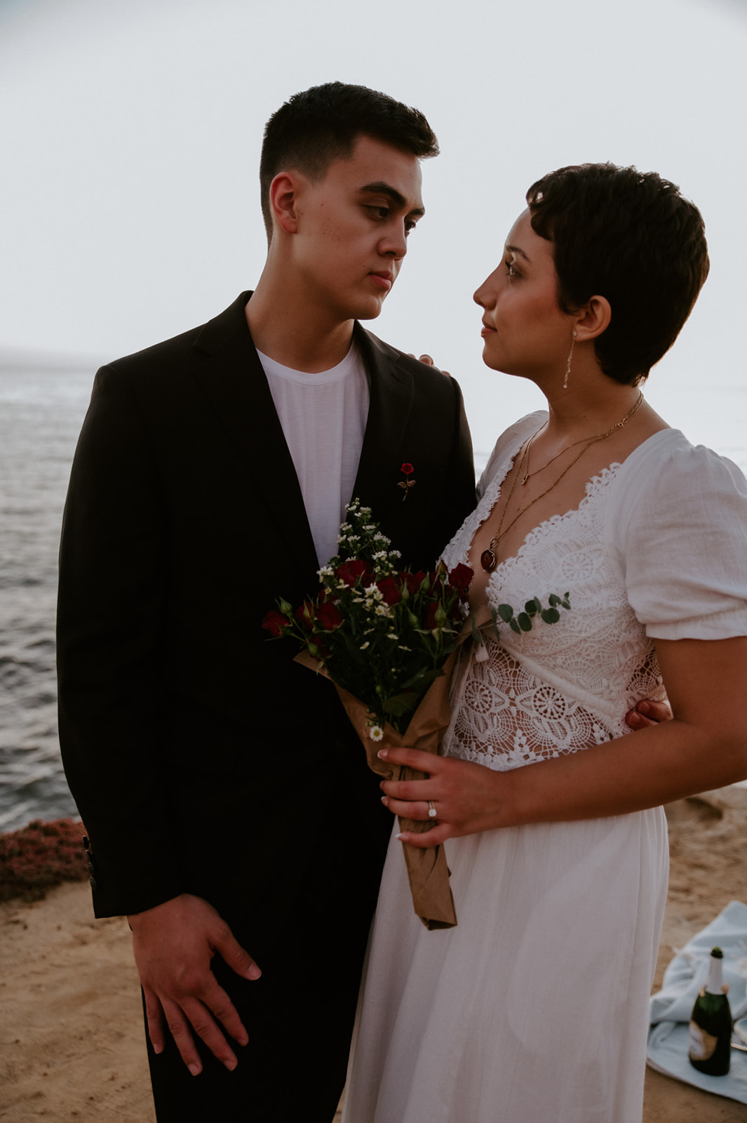 Couple holding a small bouquet and looking at each other by the ocean, a simple and meaningful detail from their elopement checklist.

