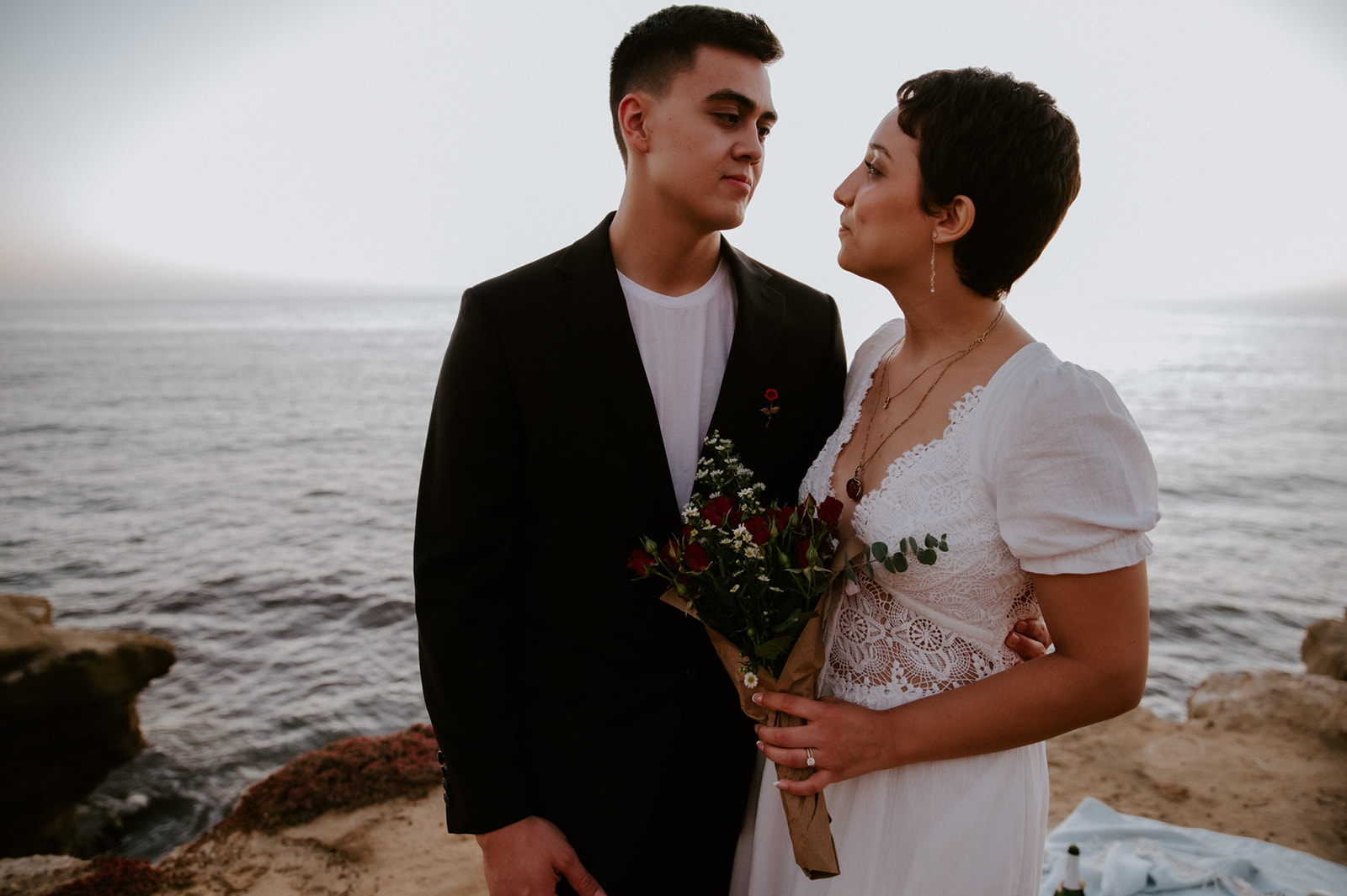 Couple standing close together holding a small bouquet on a coastal cliff, a simple and intentional elopement detail.
