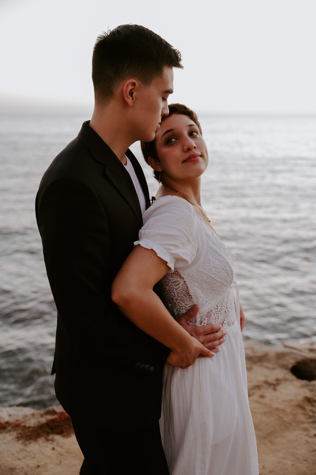 Groom kissing bride’s forehead as they stand close by the ocean, a quiet and grounding moment during their elopement day.
