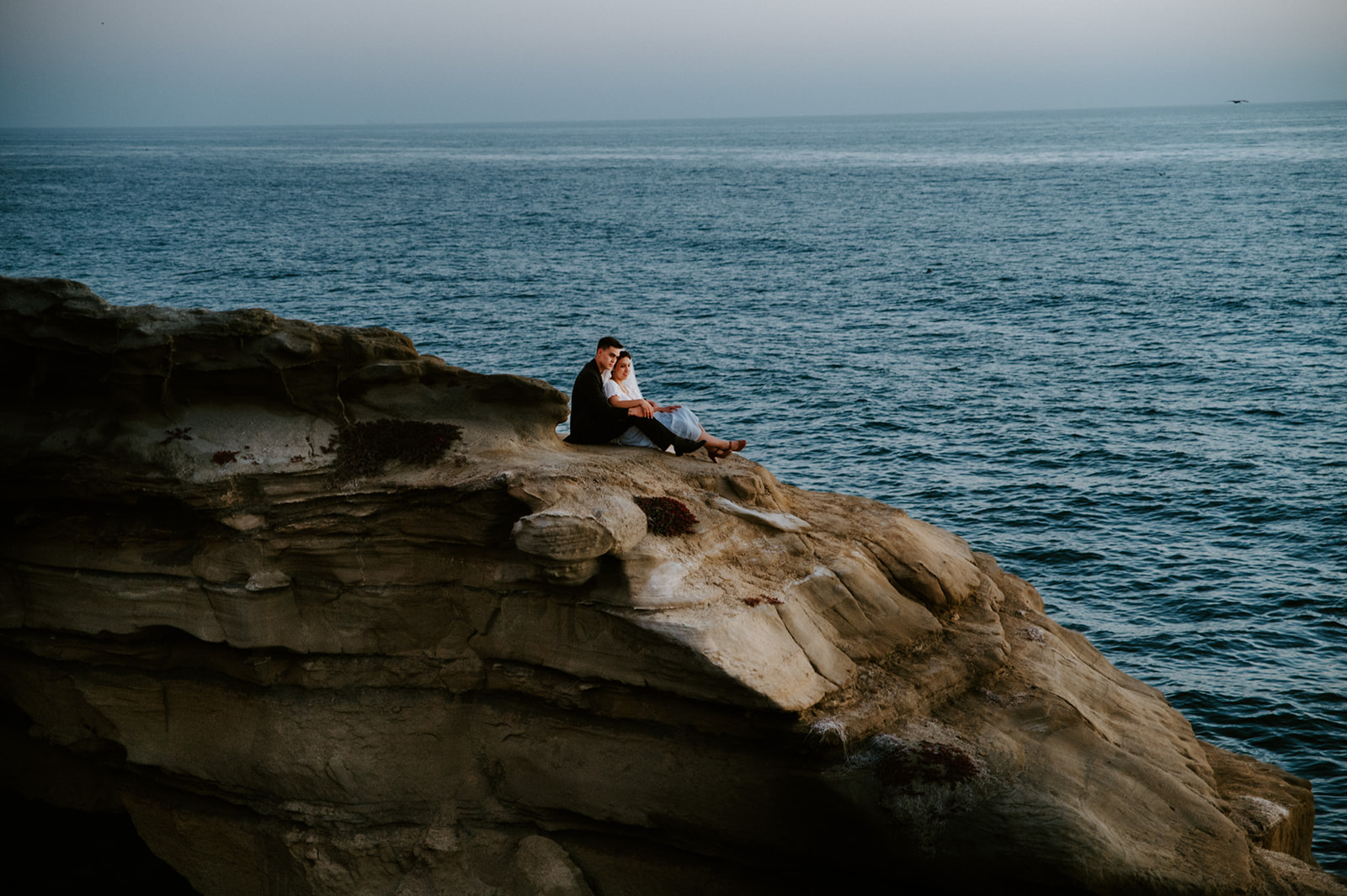 Couple sitting together on the edge of a coastal cliff overlooking the ocean, taking in the quiet after their elopement checklist day.
