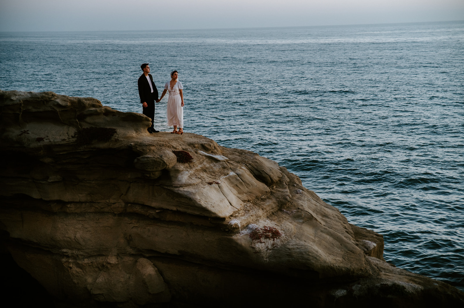 Wide shot of couple standing hand in hand on a rugged coastal cliff above the ocean, an epic landscape moment from their elopement checklist.
