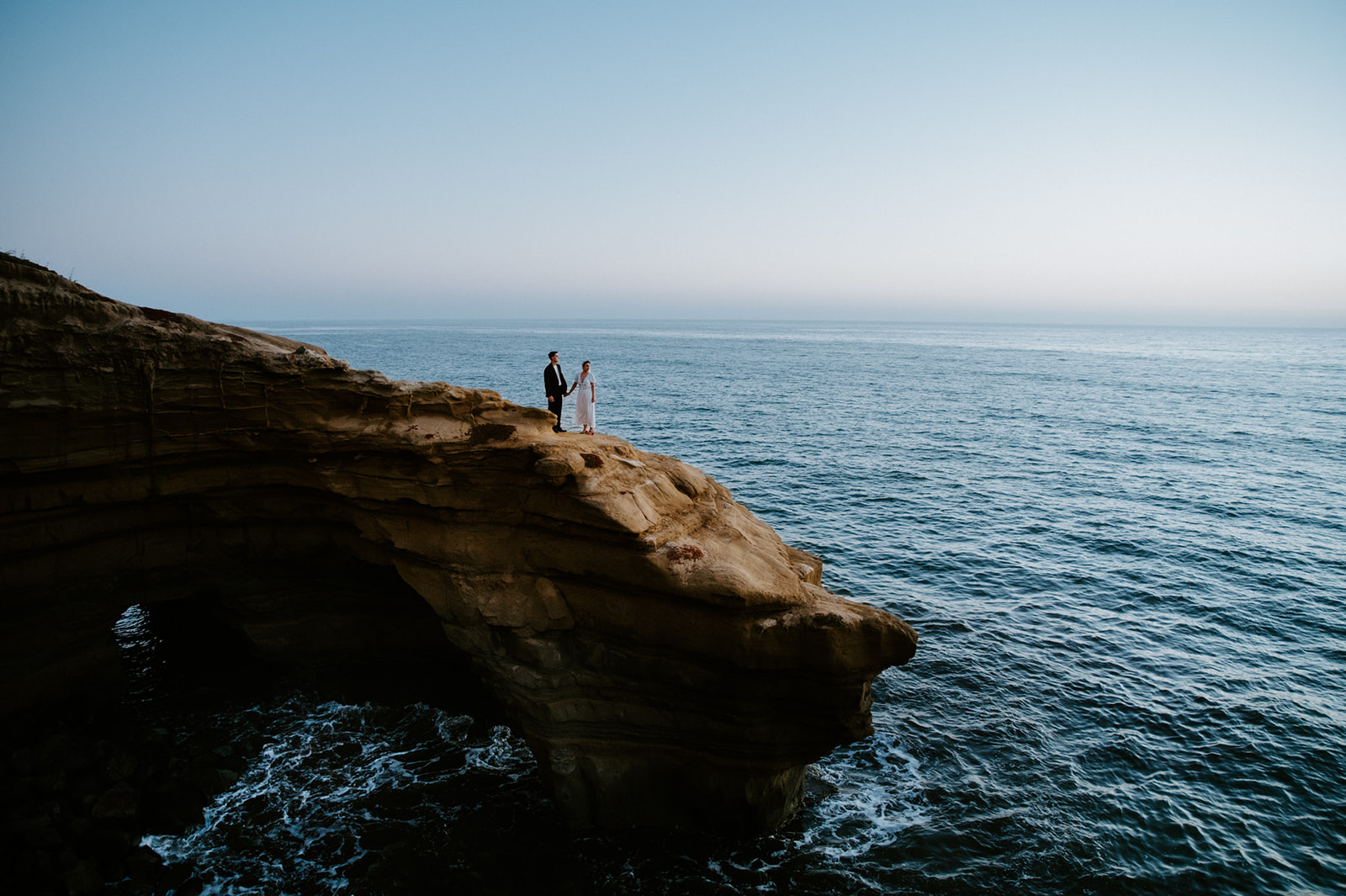 Wide coastal cliff scene with couple standing hand in hand above the ocean, an epic landscape moment to include in your elopement checklist.
