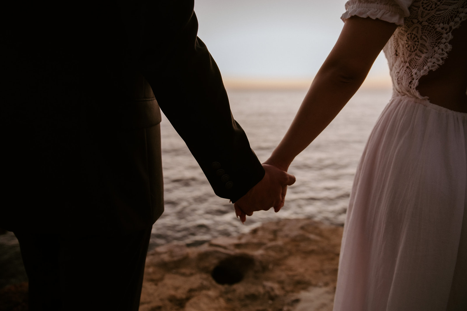 Close-up of couple holding hands near the ocean at golden hour, a grounding moment that reflects the heart of an elopement checklist.
