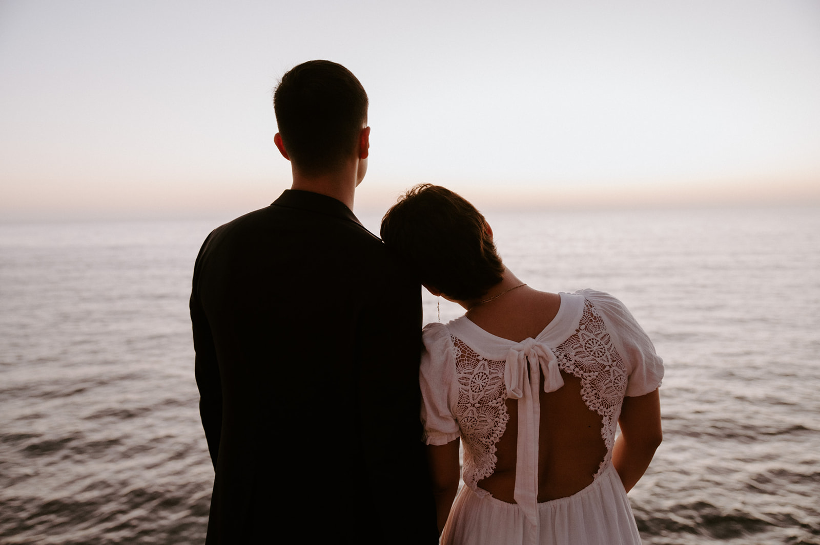 Couple standing side by side facing the ocean, the bride resting her head on her partner’s shoulder during their elopement.
