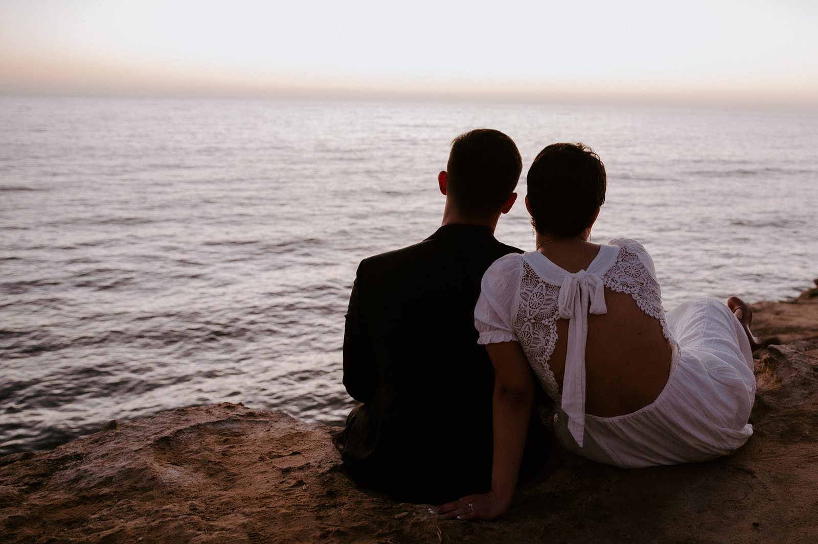 Couple sitting side by side on a cliff overlooking the water at sunset, a quiet pause built into their elopement checklist.
