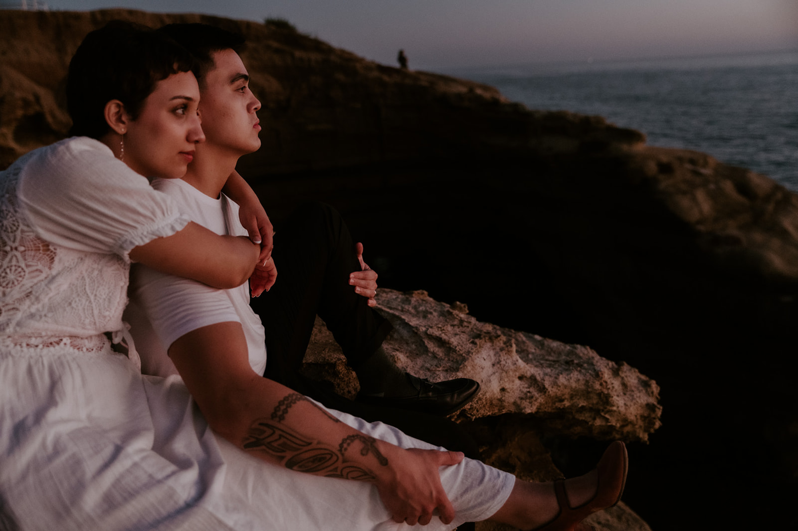 Couple sitting together on a rocky overlook at dusk, wrapped up in each other while watching the coastline fade into evening light.
